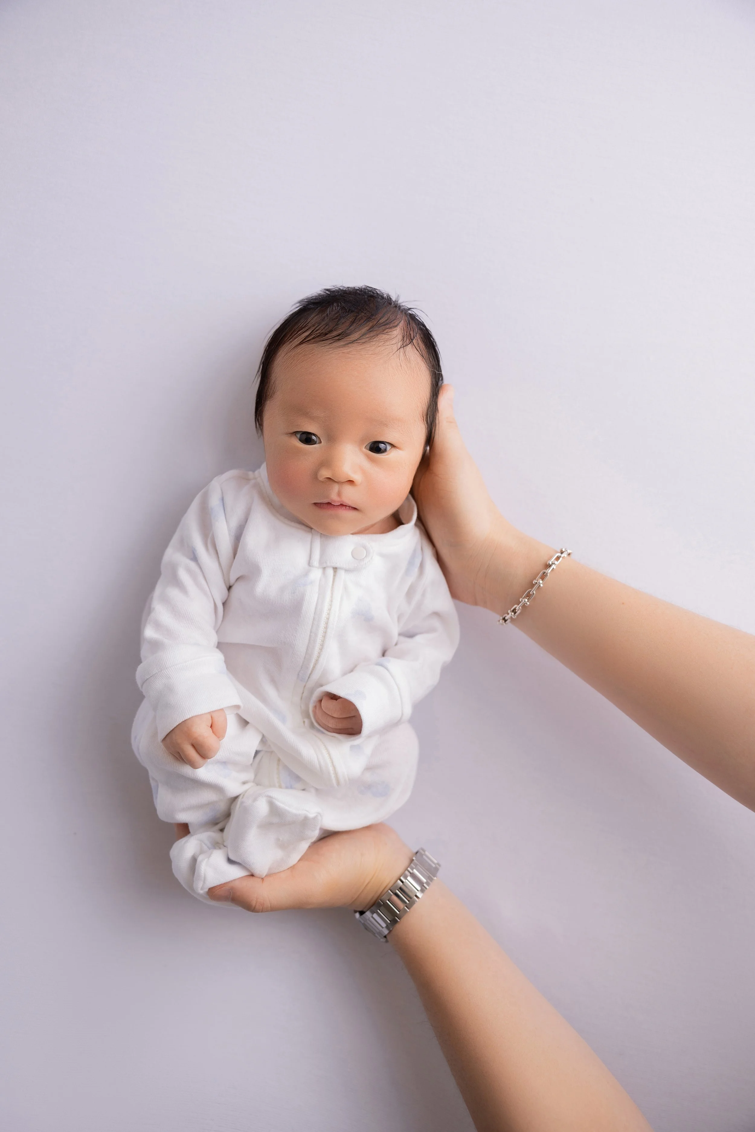 A newborn baby in a white onesie held gently in adult hands against a plain white background.