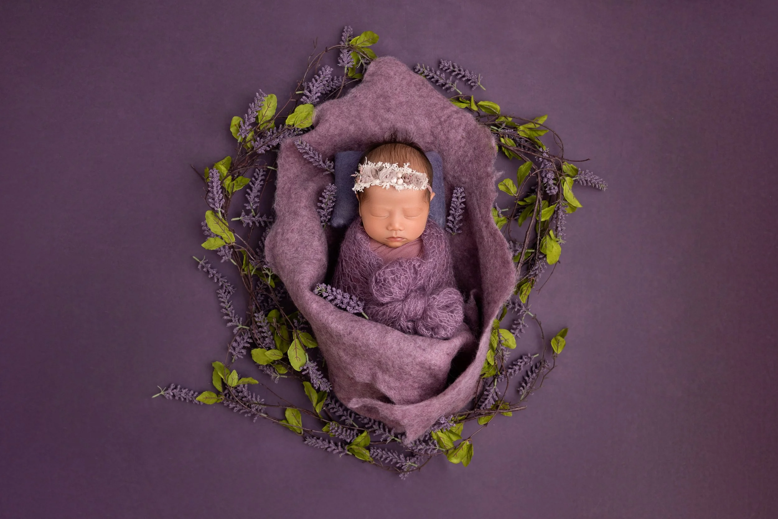 A sleeping baby girl wrapped in purple fabric, wearing a floral headband, surrounded by a lavender-colored blanket and a border of green leaves and lavender sprigs on a purple background.