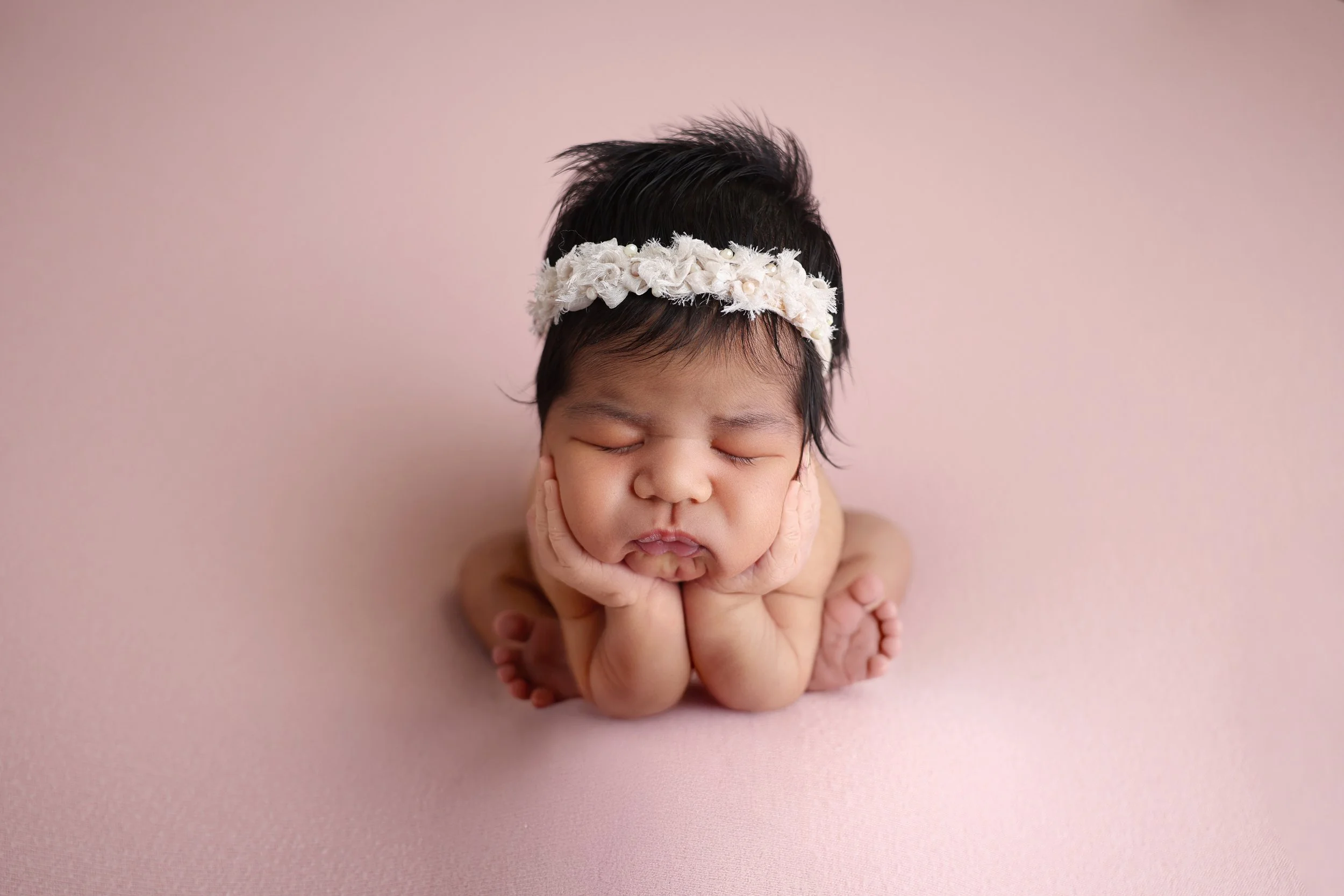 A sleeping baby with a floral headband, resting on a soft pink surface with hands under cheeks.