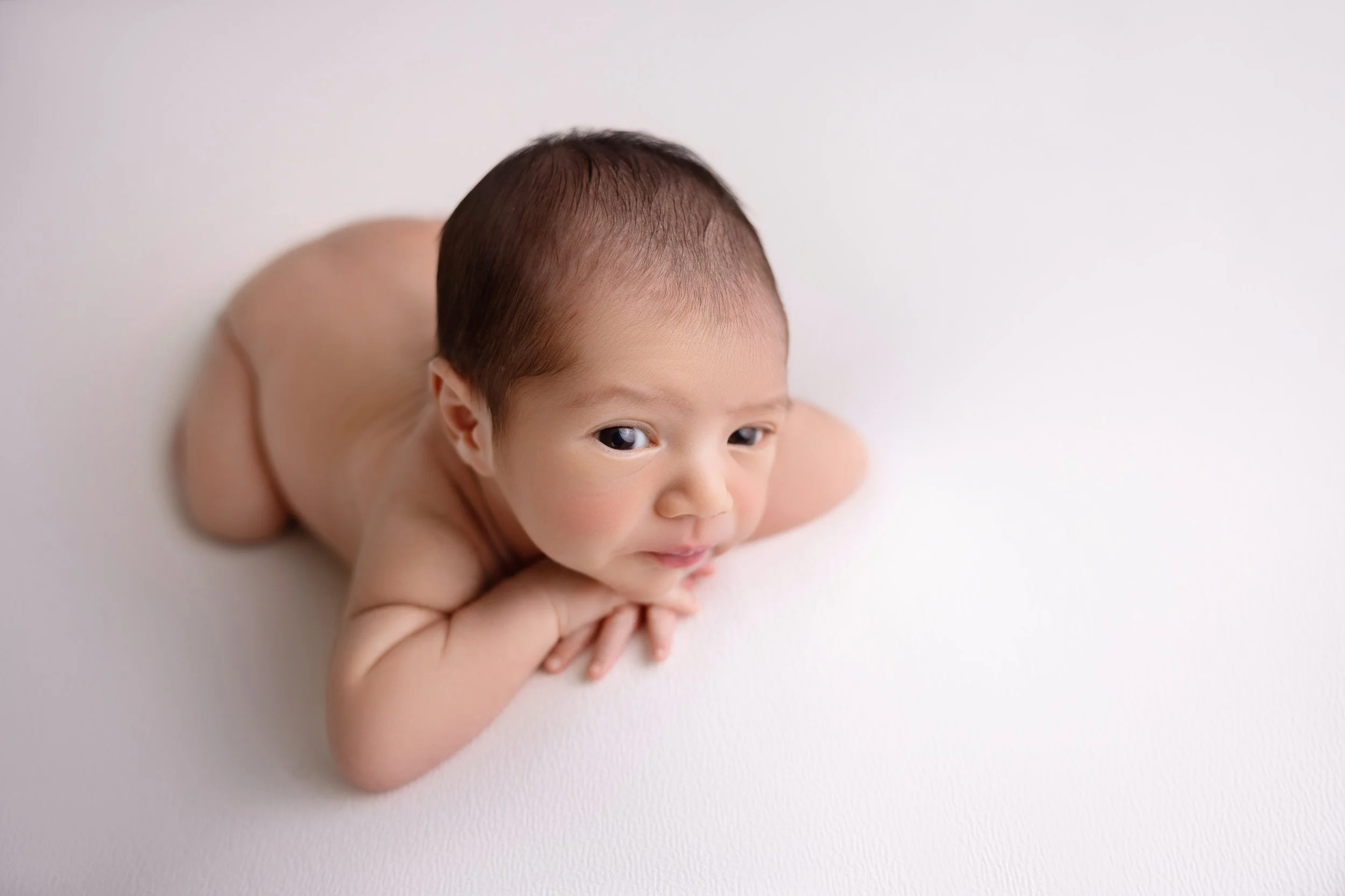 A baby lying on a white surface with their head resting on their arms, looking at the camera with a gentle smile.