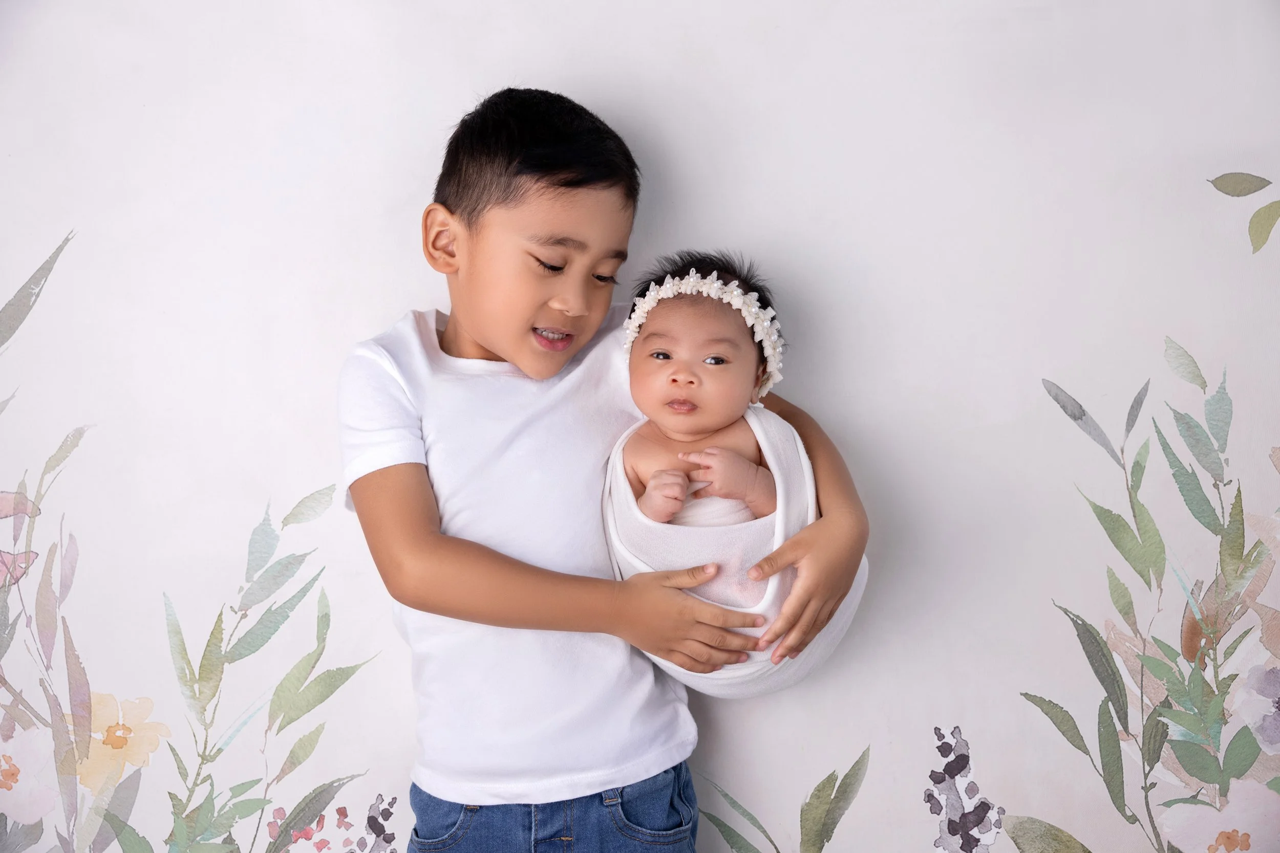 A young boy holding a baby girl wrapped in a white blanket against a floral backdrop.