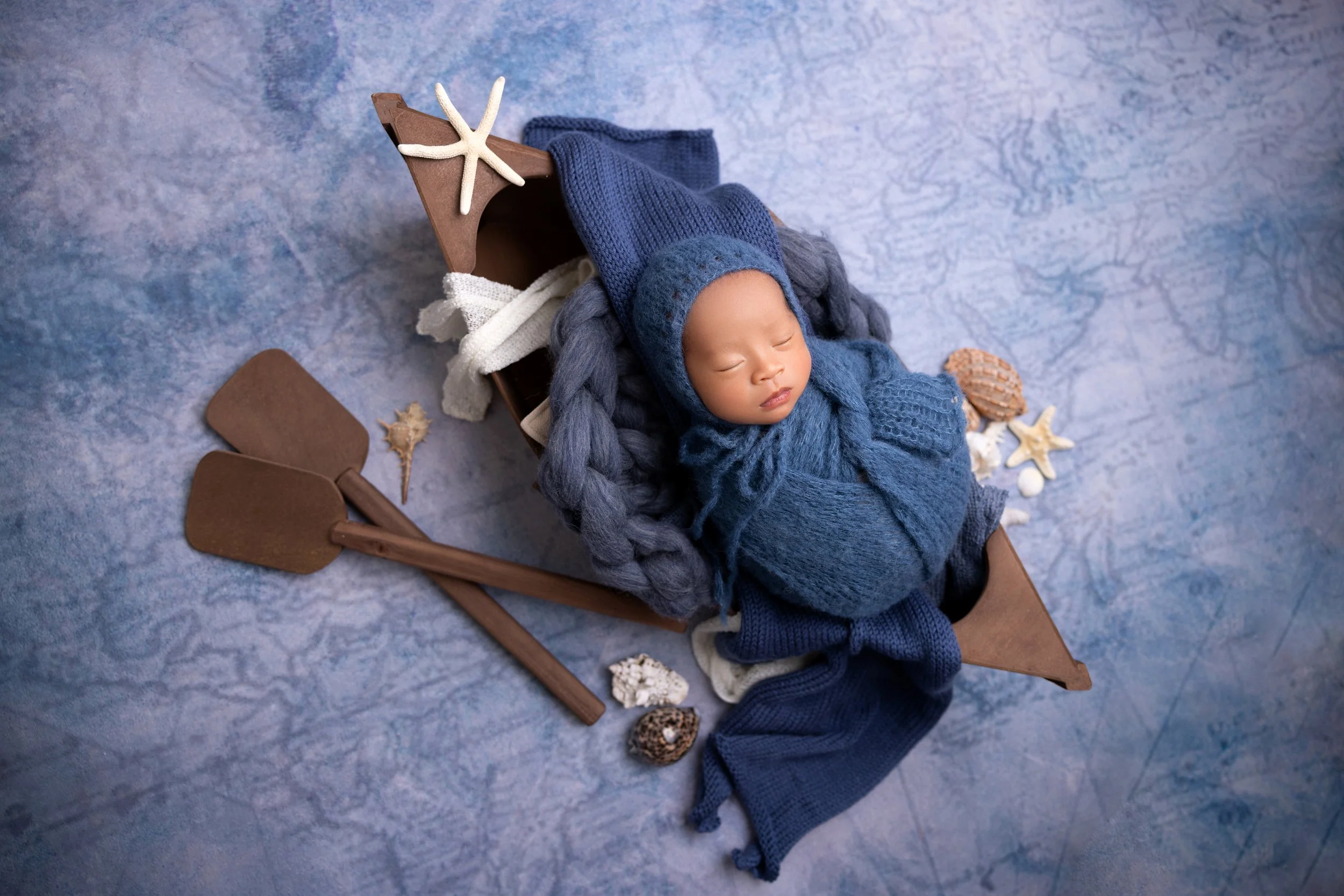 A sleeping baby dressed in blue, wrapped in a knit blanket and wearing a bonnet, lying in a wooden boat with seashells, starfish, and gardening tools around him, on a blue textured surface.