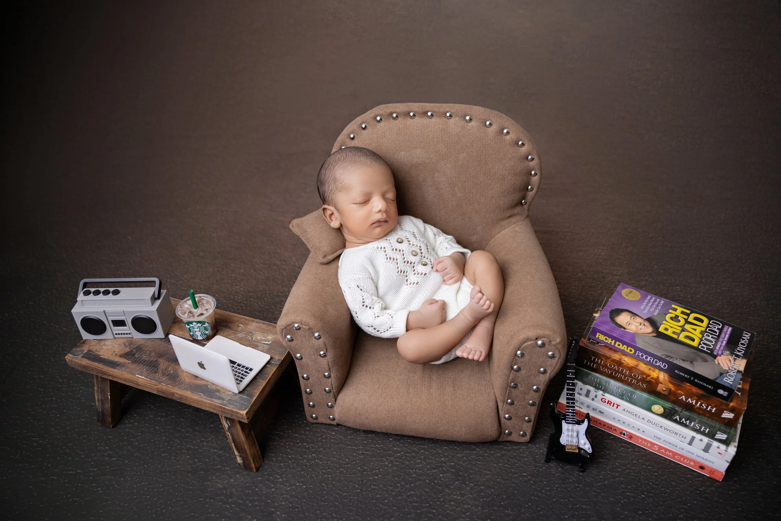 A sleeping baby in a small armchair with a wooden side table, a Starbucks drink, a mini laptop, a boombox, a guitar, and a stack of books beside them.