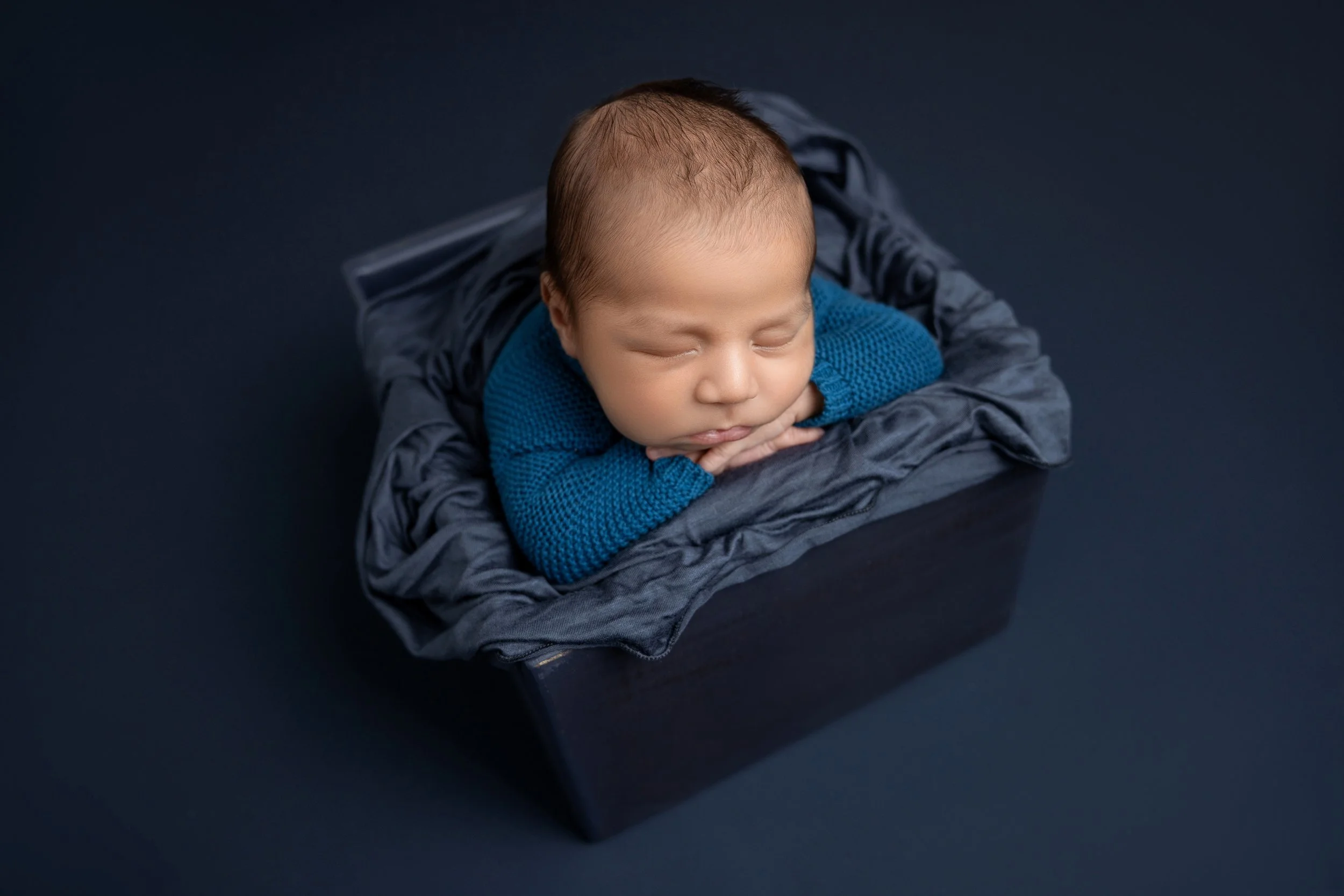 A sleeping baby in a dark blue box, resting on gray cloth, wearing a blue knitted sweater.
