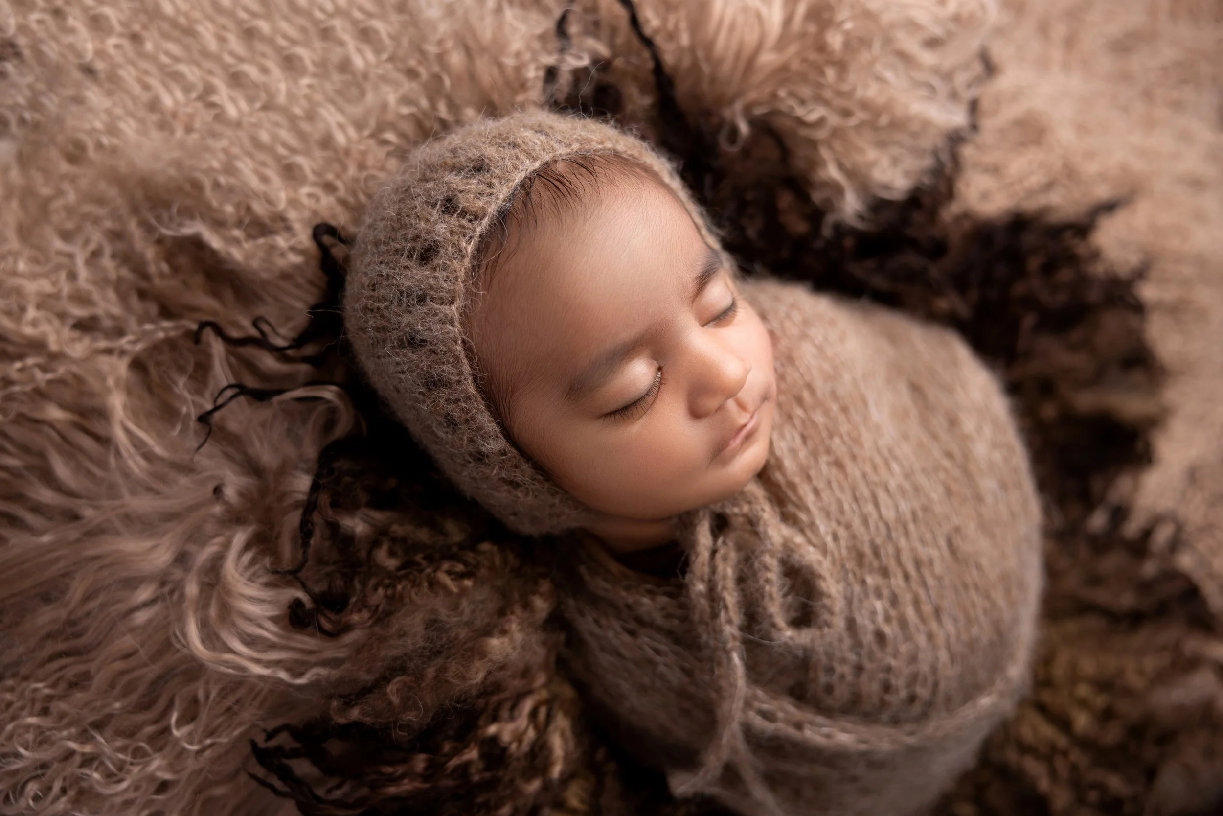 A baby dressed in a knitted brown outfit and hat, peacefully sleeping on a soft, brown, furry surface.
