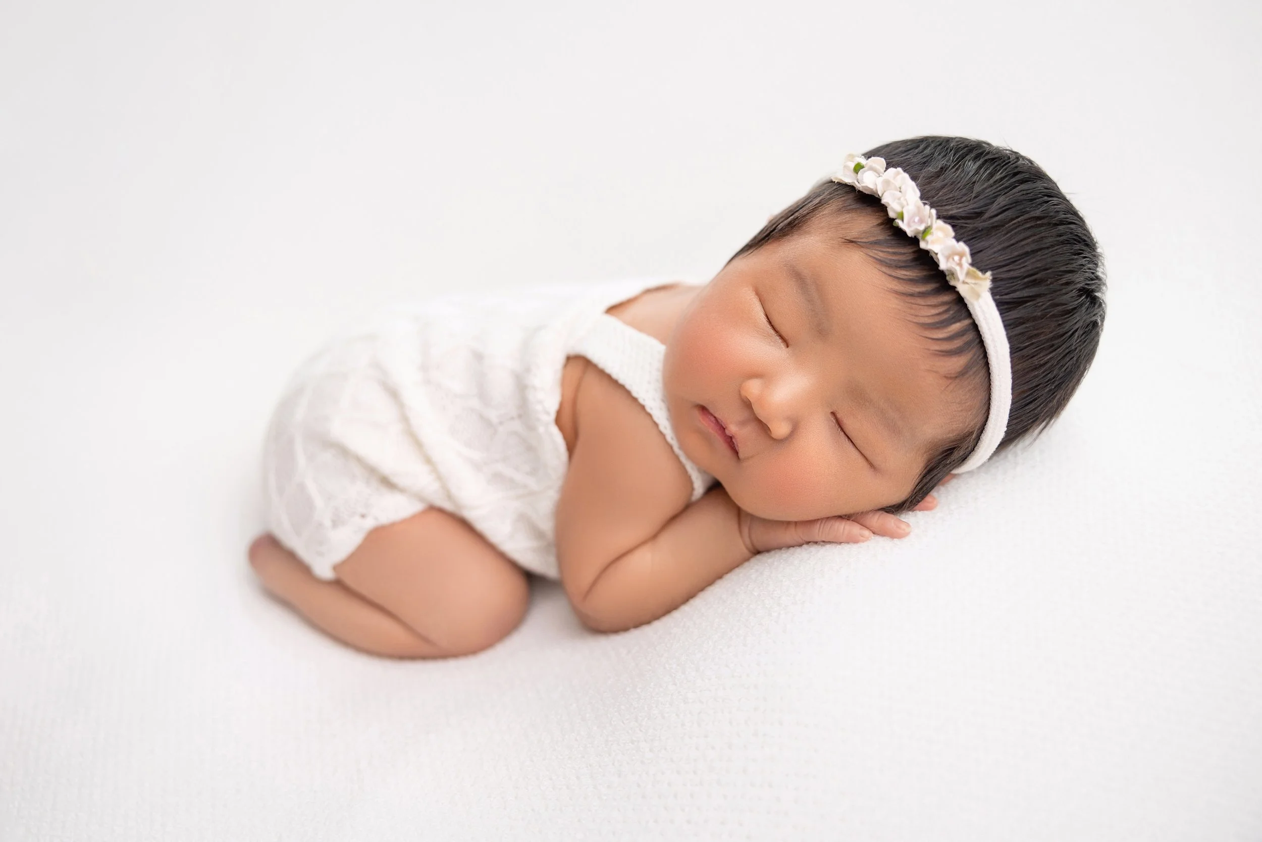 A sleeping baby girl with short black hair wearing a white headband with flowers, lying on her side with hands under her cheek on a white surface.