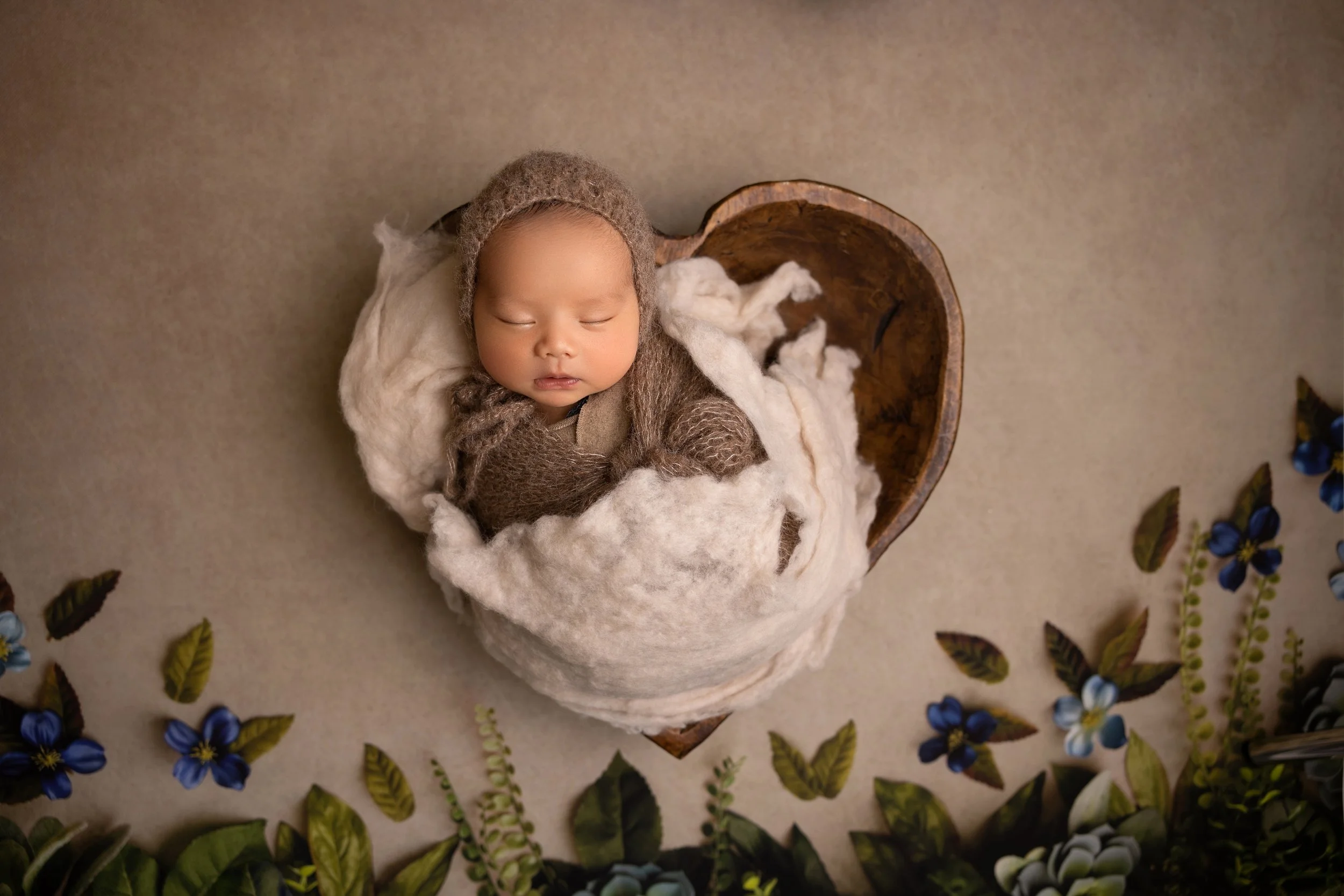 A sleeping baby wearing a brown knit outfit and bonnet, resting in a wooden bowl on a beige carpet, surrounded by blue flowers and green leaves.