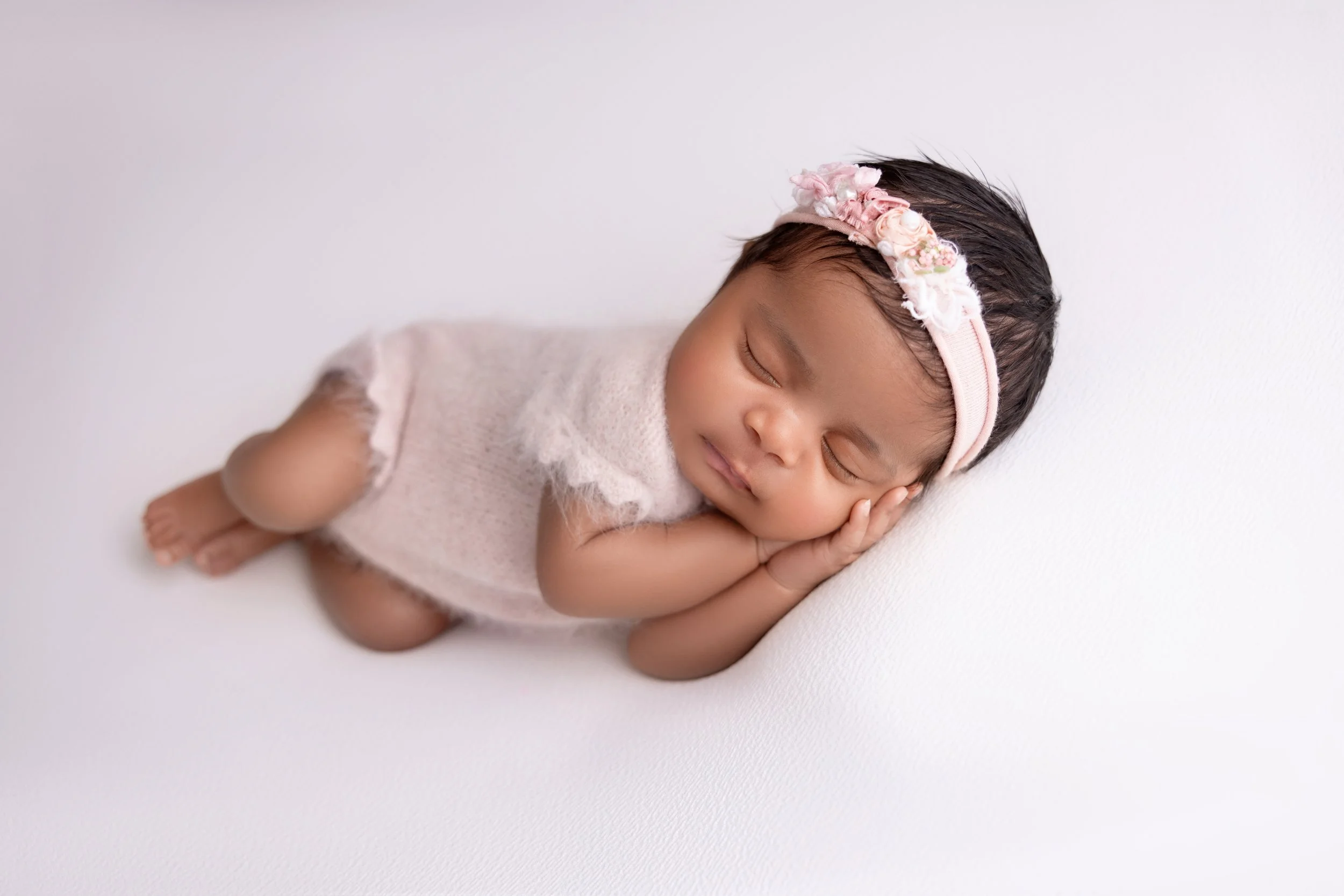 A sleeping baby girl with a floral headband, lying on her side with her hands clasped under her cheek, against a white background.