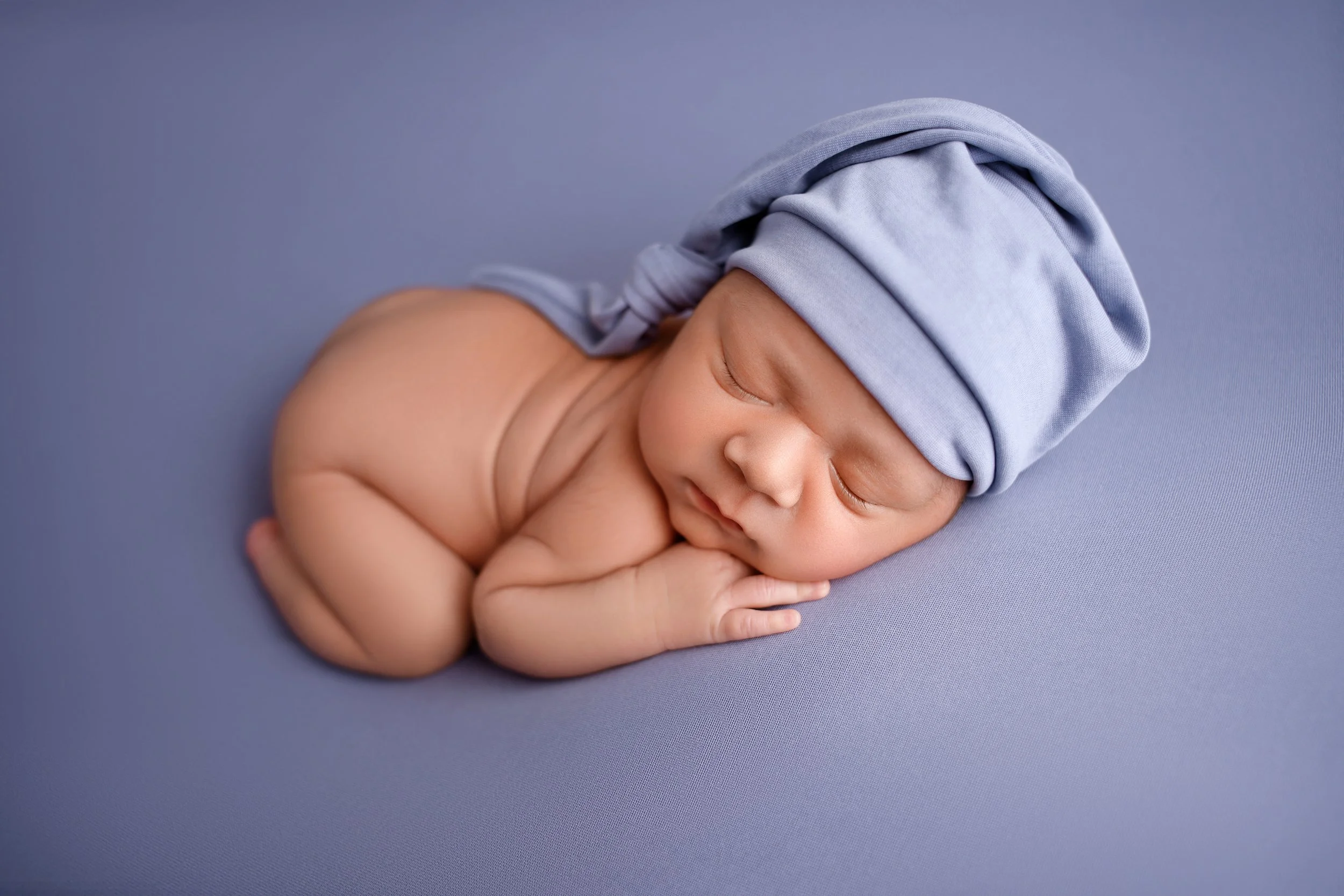 A sleeping newborn baby curled up on a soft blue surface, wearing a matching blue hat and resting peacefully.