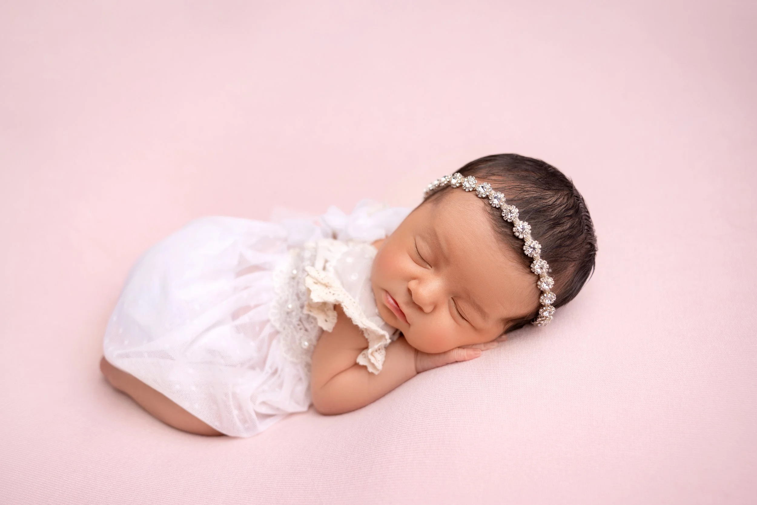 Cute sleeping newborn girl wearing a white dress and a sparkling floral headband, lying on her side on a soft pink surface