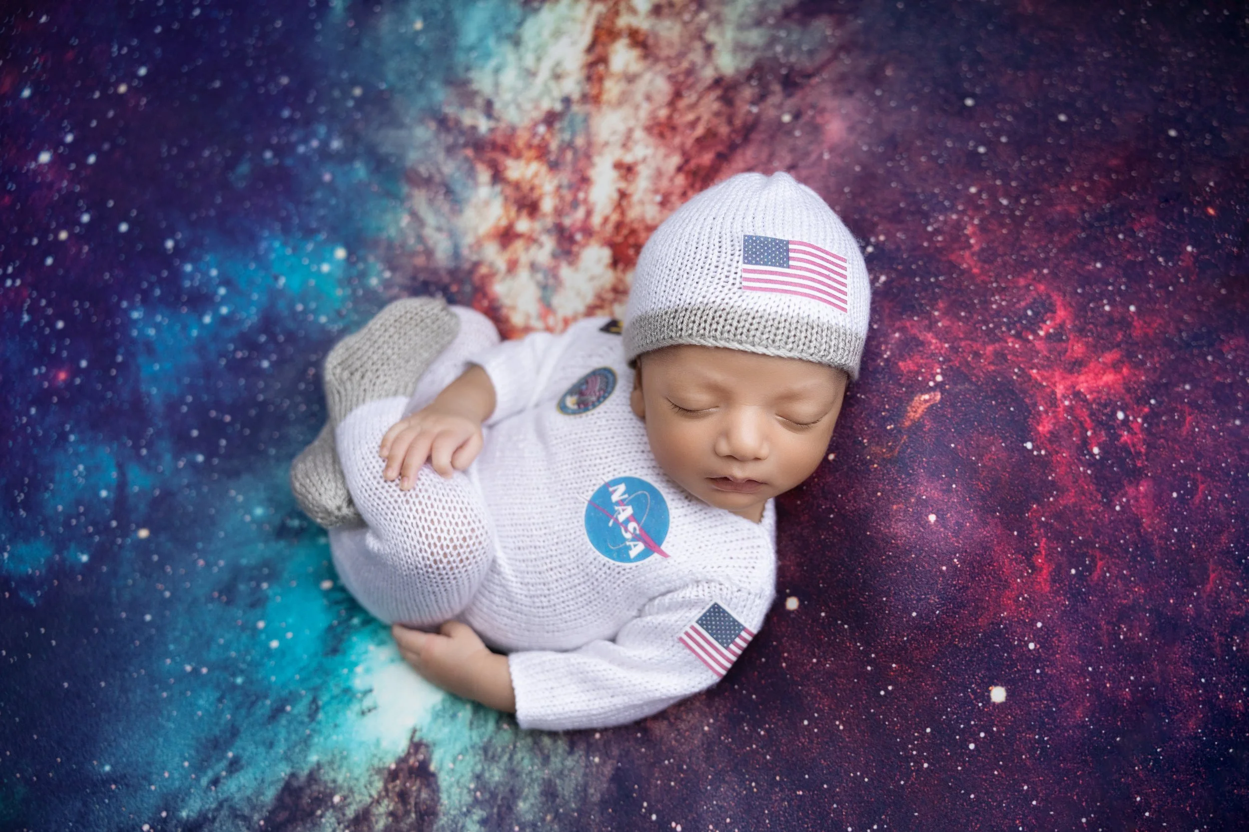 A sleeping baby dressed in a white NASA spacesuit and a cap with an American flag, lying on a colorful, galaxy-themed blanket.
