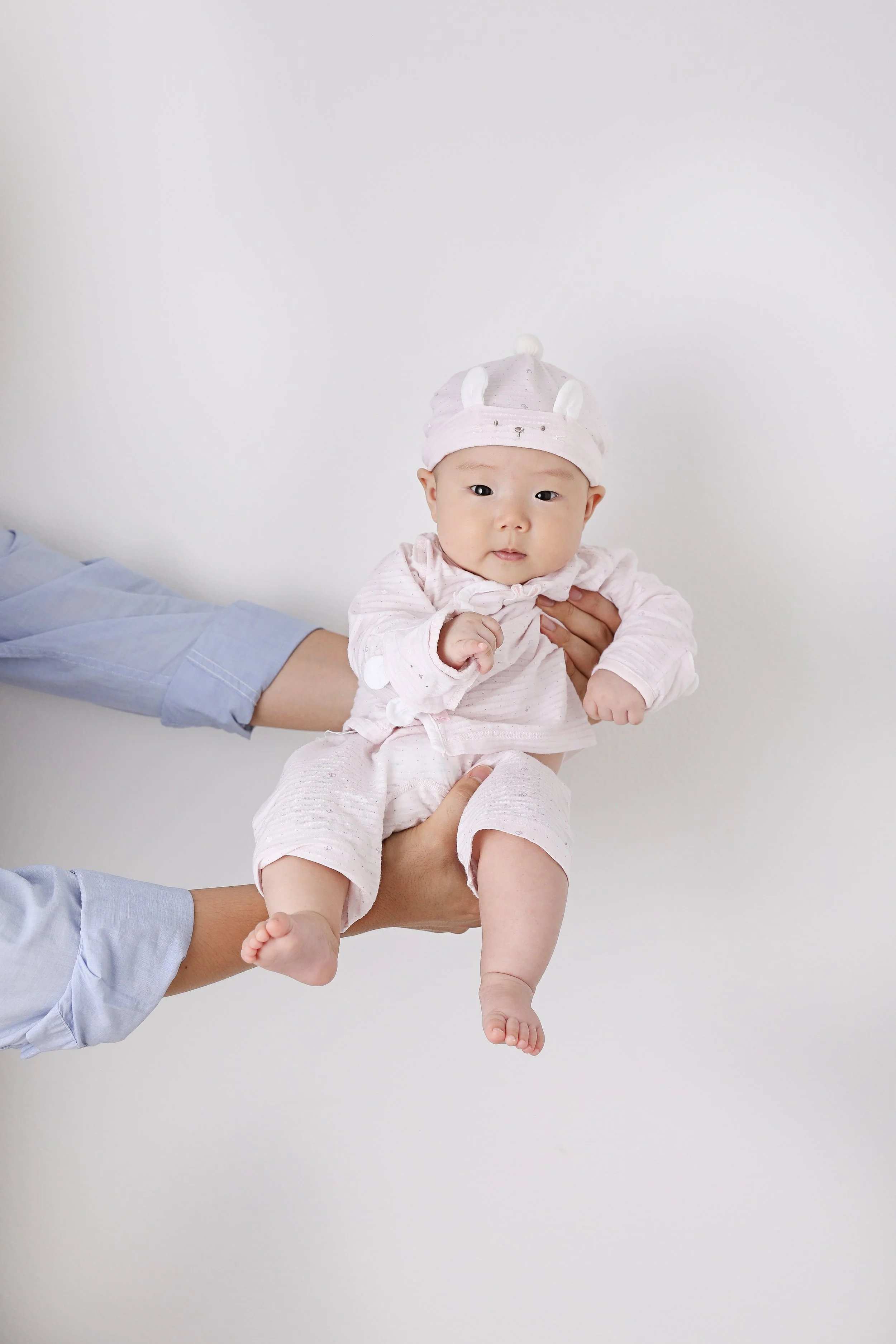 A baby dressed in pink clothing and a pink hat with white animal ears, being held in the air by two people against a plain white background.