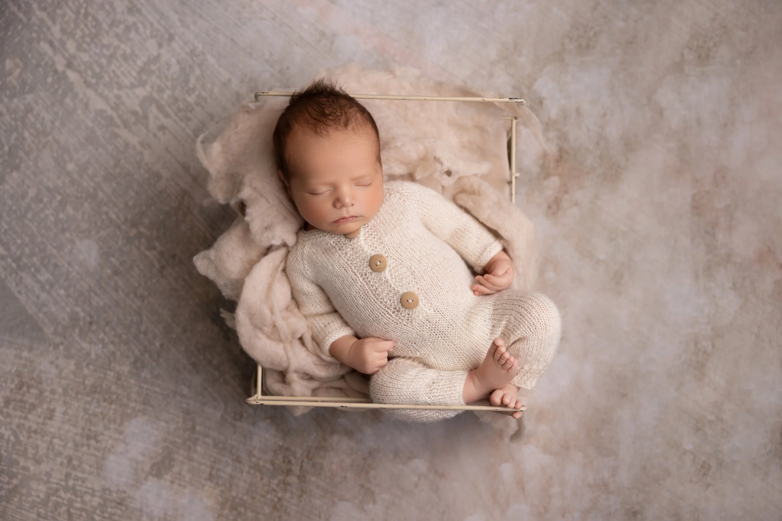 A sleeping baby dressed in a cream-colored knitted outfit lying in a small metal bed with soft blankets on a textured surface.