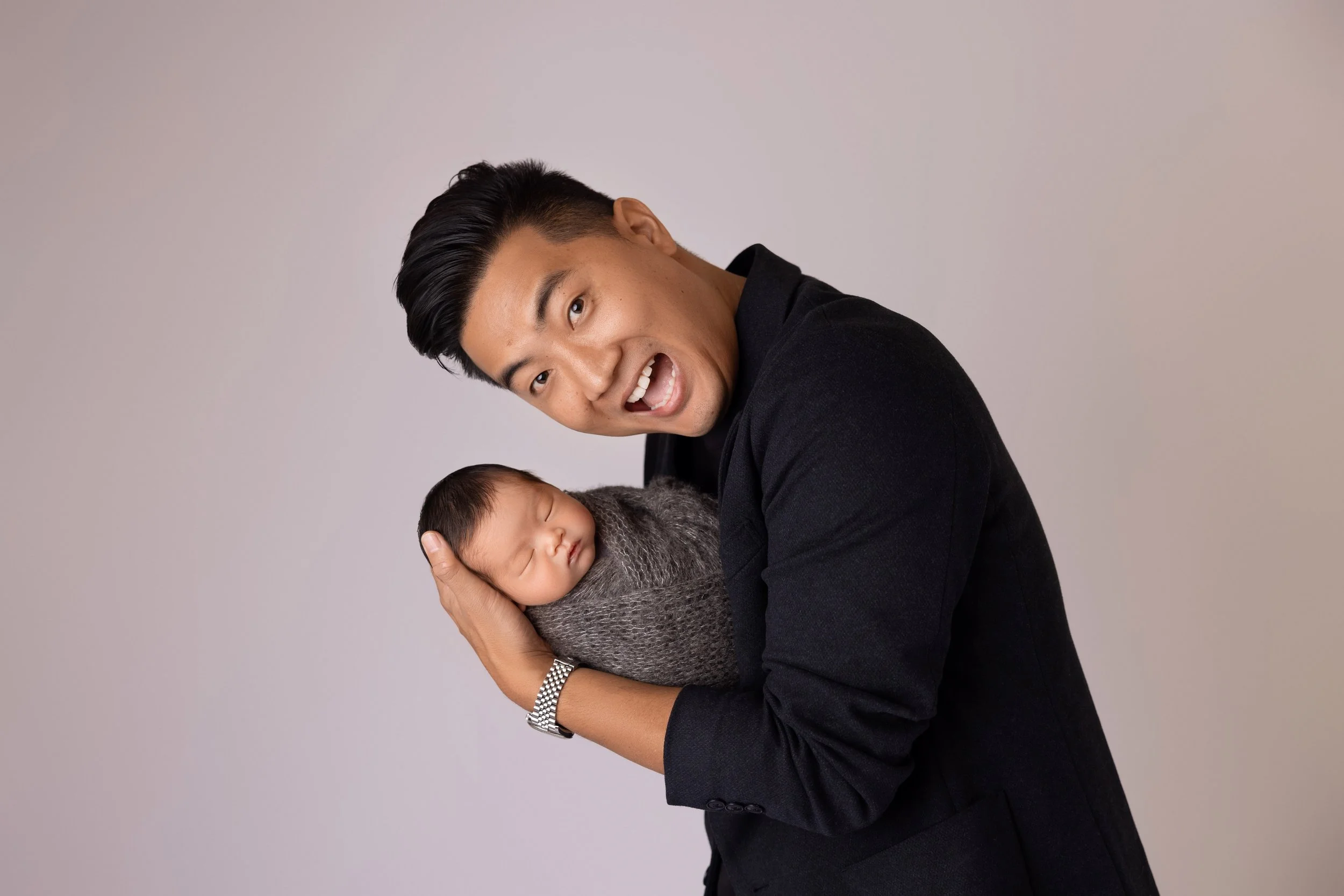 A young man in a black suit holding a sleeping newborn wrapped in gray knit blanket, against a plain light background.