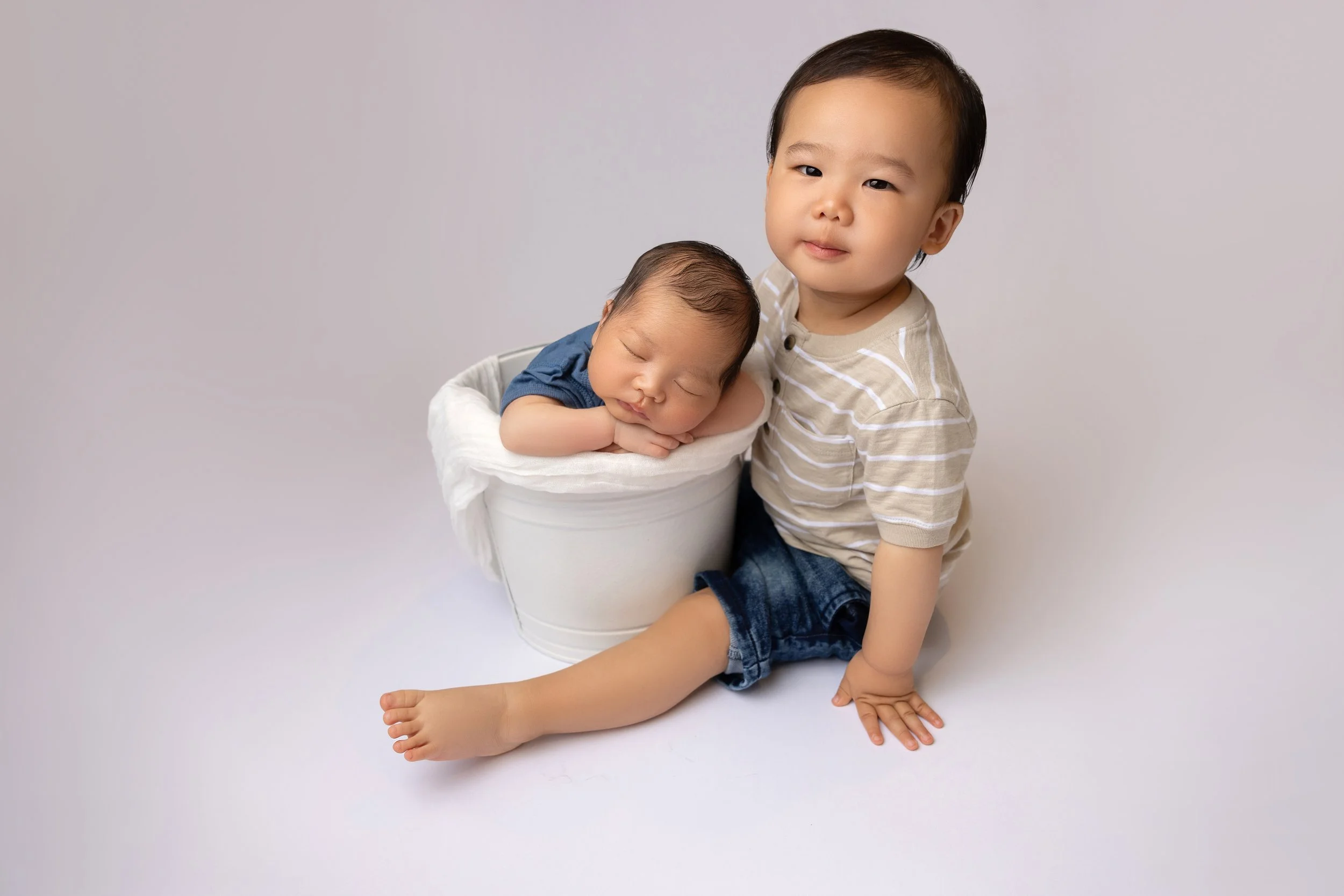 A young boy sitting on the floor next to a bucket with a sleeping baby inside, both children have dark hair and are dressed casually, against a neutral background.