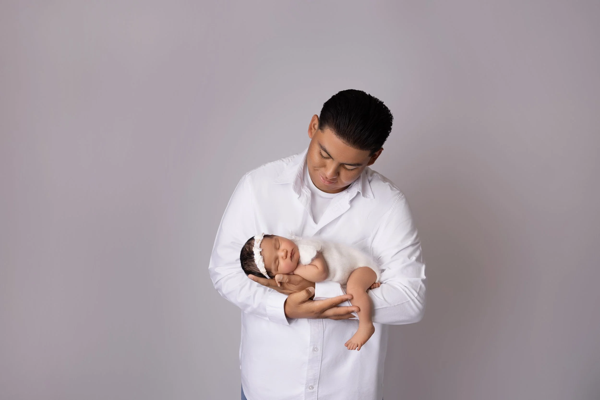 A man in a white shirt holding a sleeping newborn baby with a white headband.