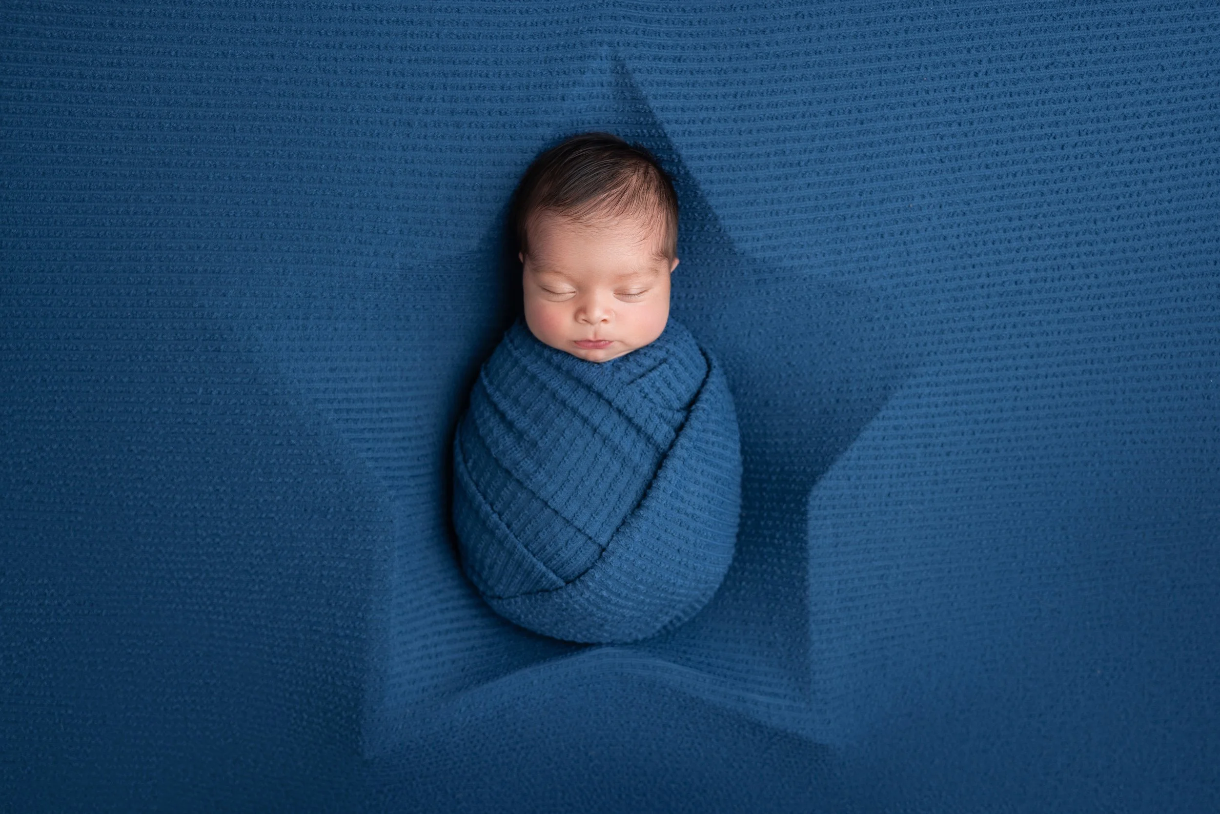 A newborn baby sleeping, wrapped in a matching blue knit blanket, lying on a blue textured surface.