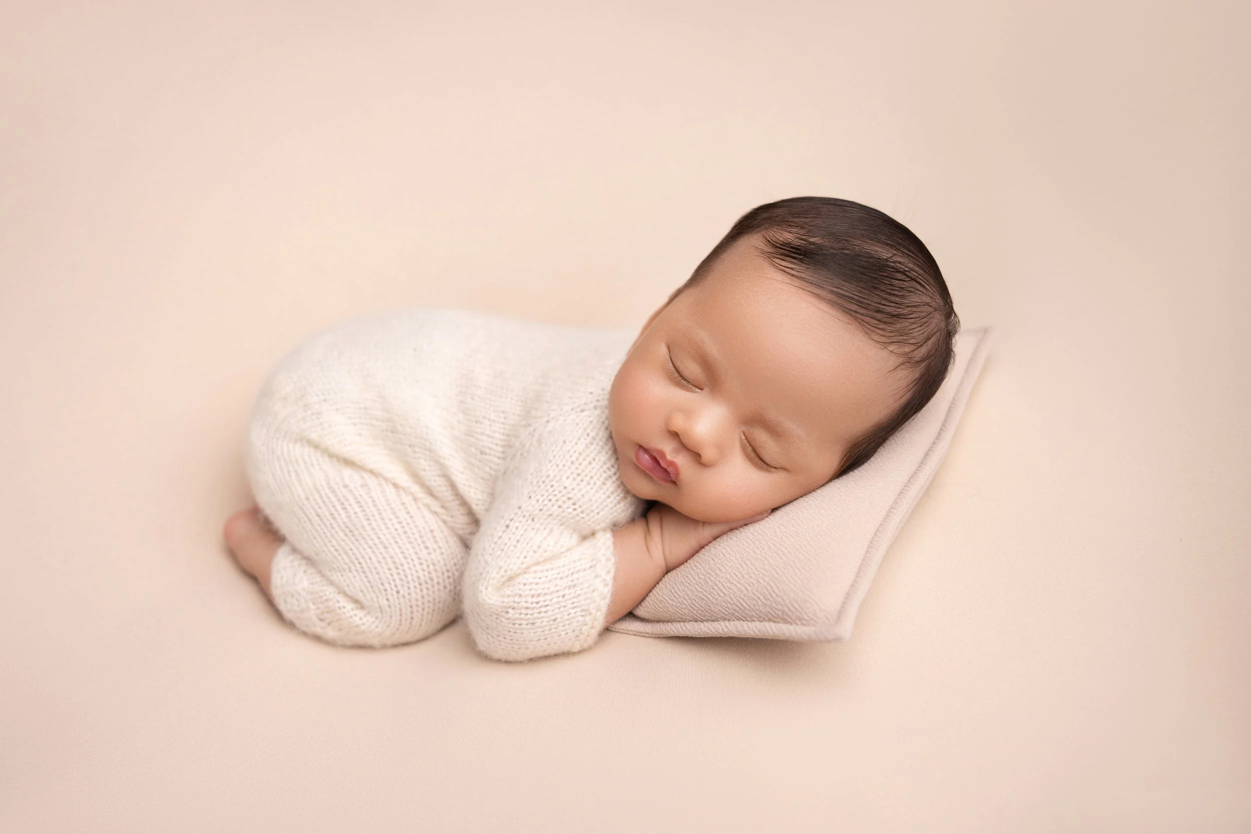 A sleeping infant with light skin and dark hair lying on a small pillow on a beige background.