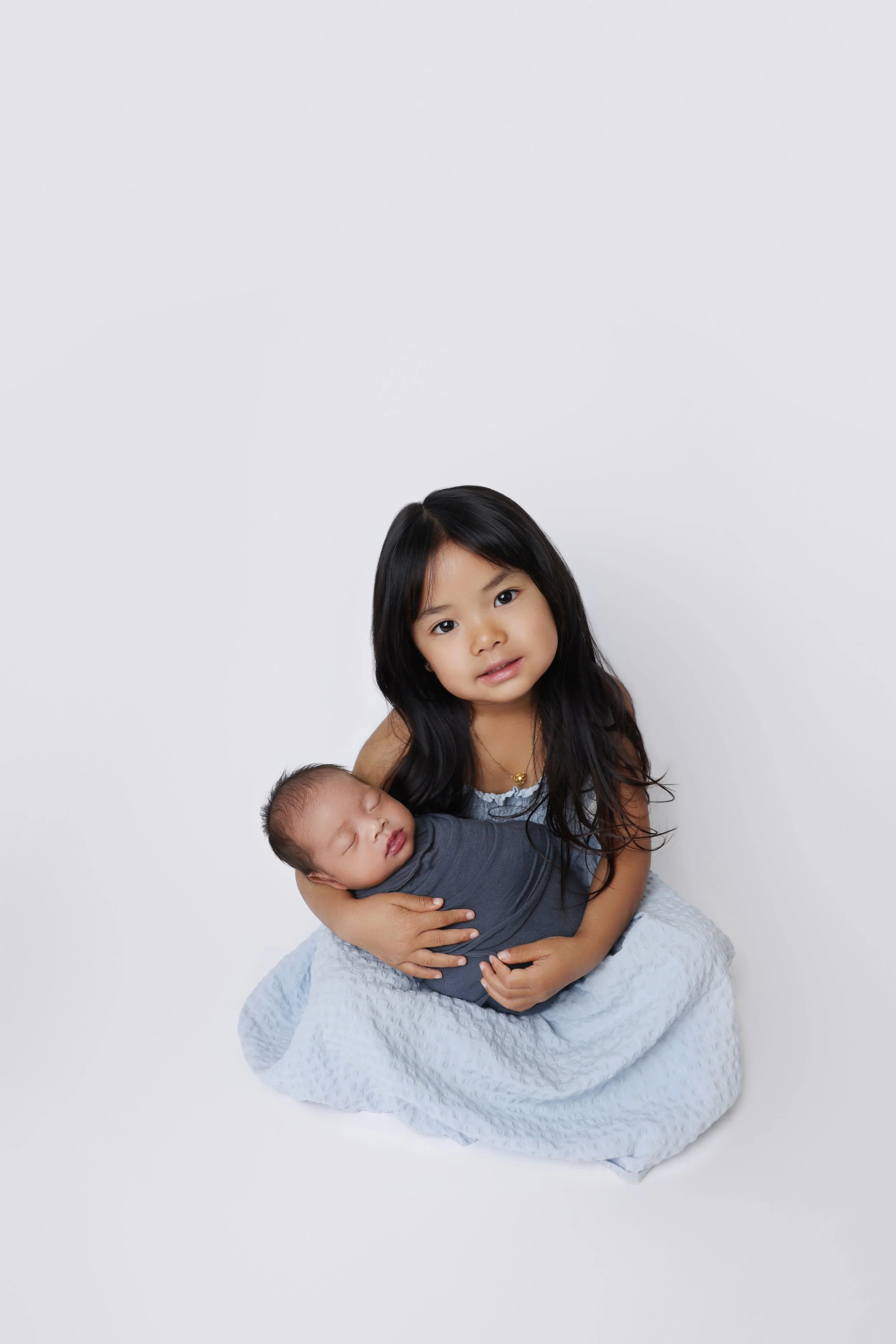 A young girl with long black hair gently holding a sleeping baby in a blue outfit, sitting on a white surface against a plain white background.