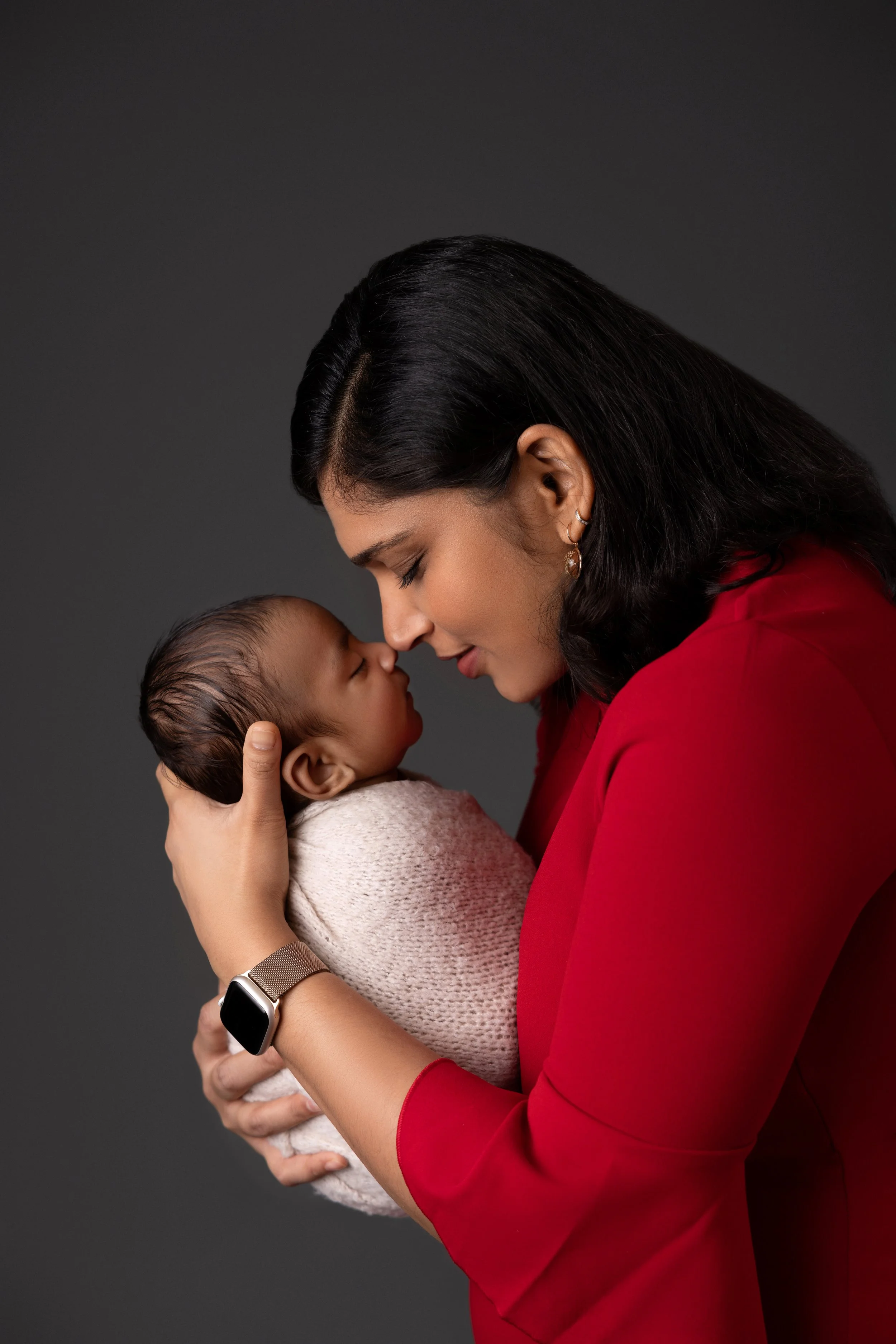 A woman holding a sleeping newborn baby close, their foreheads touching, against a dark gray background.