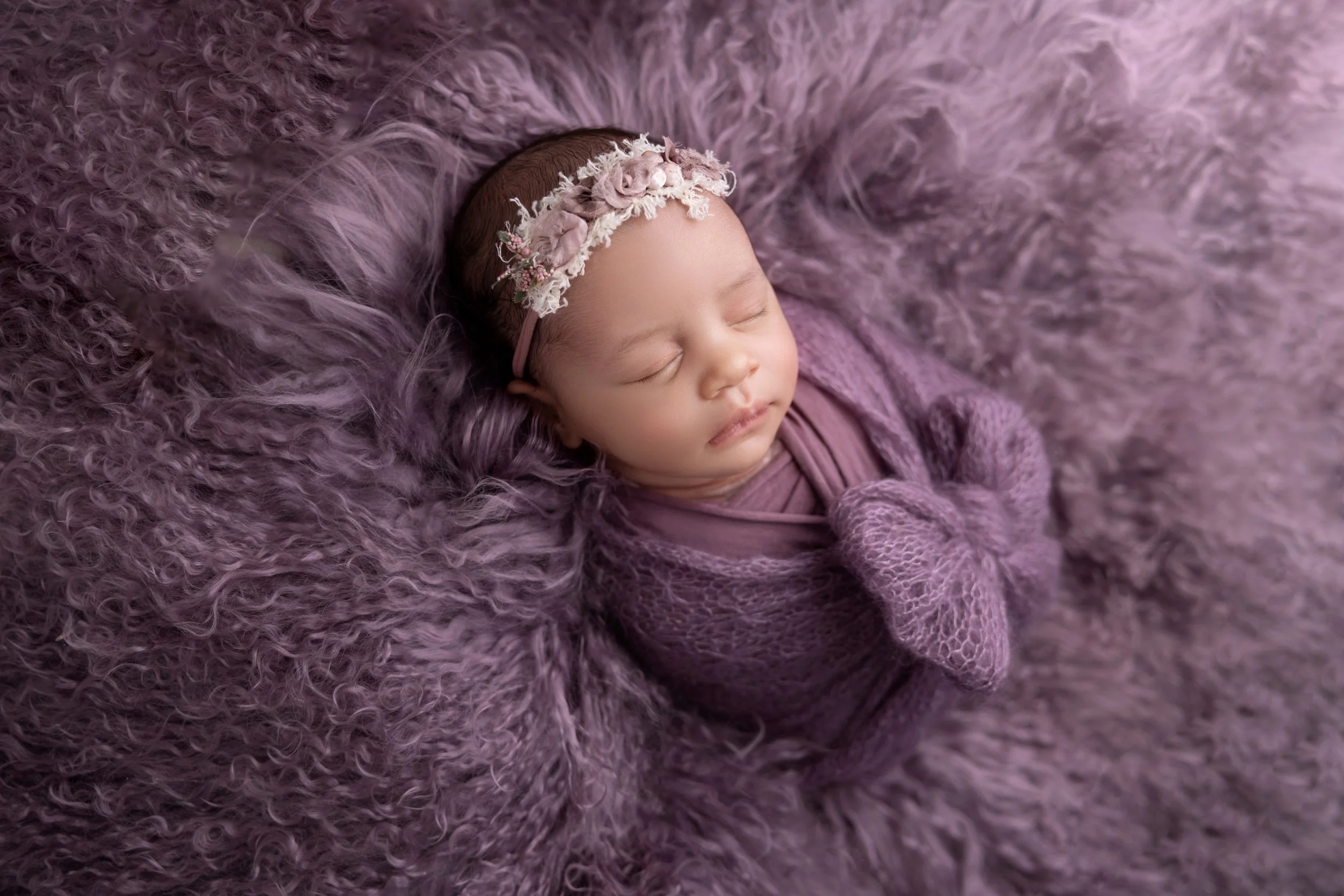 A sleeping baby wrapped in purple blanket and surrounded by purple furry blanket, wearing a floral headband and a purple bow on the blanket.