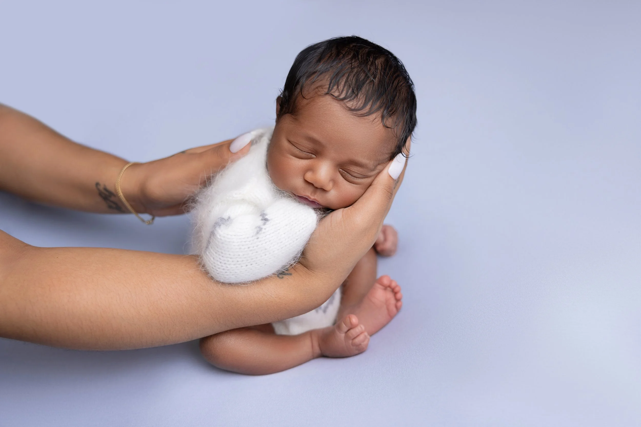 A newborn baby sleeping on a person's hand, wearing a white knit outfit, with a light blue background.