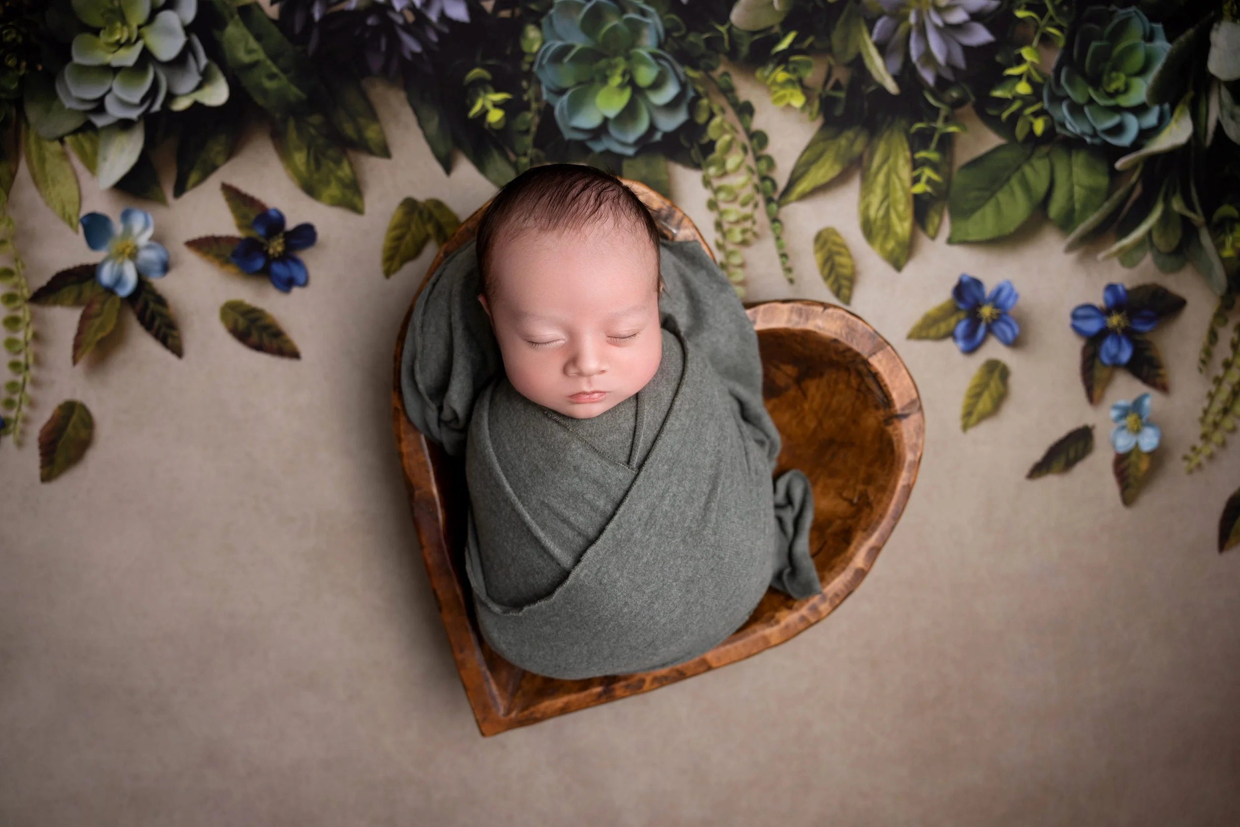 A sleeping baby wrapped in gray swaddle, lying in a wooden heart-shaped basket, surrounded by green foliage and blue flowers.