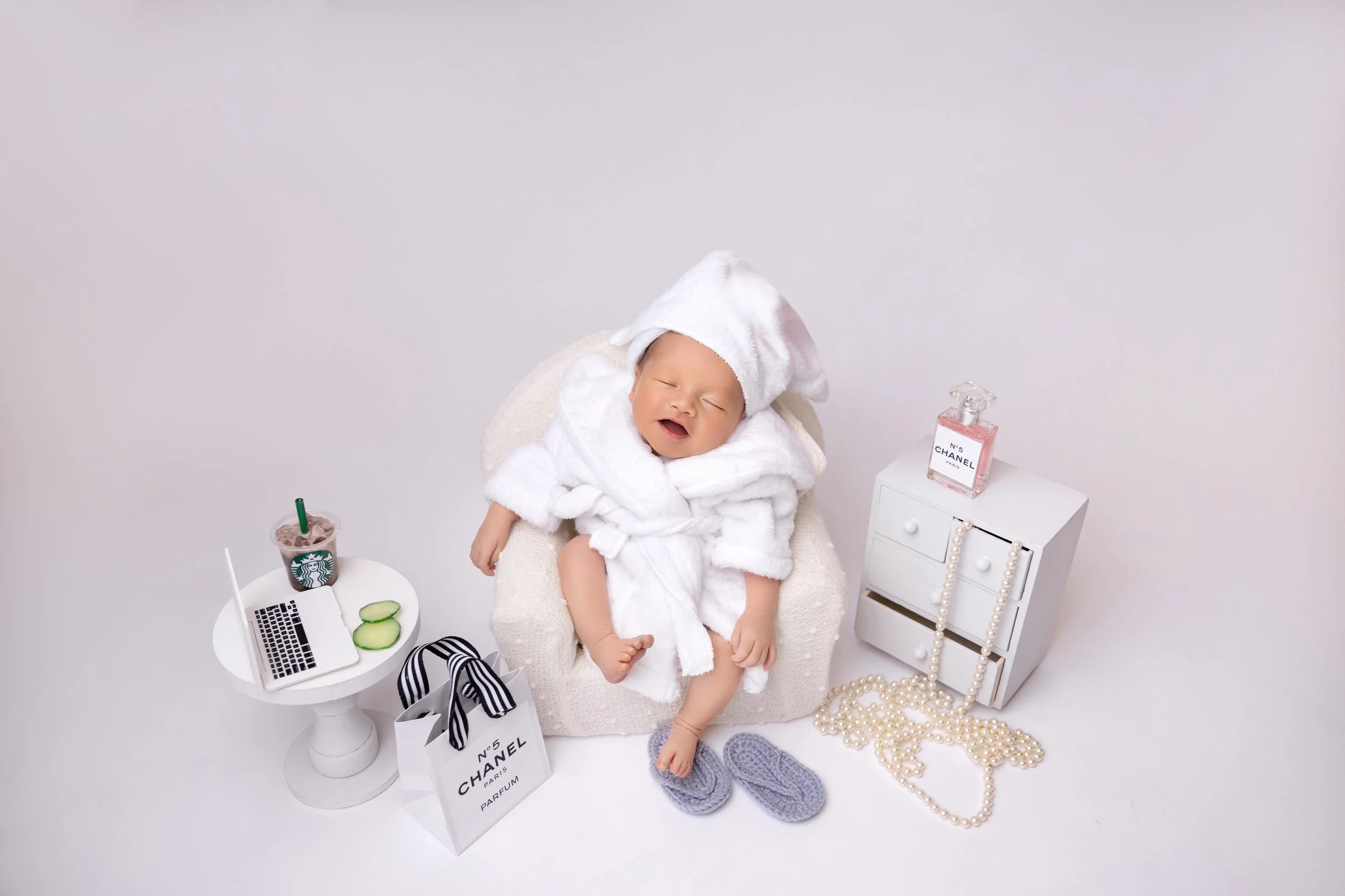A baby in a white bathrobe and a towel hat, sitting on a small armchair, surrounded by luxury accessories including a Chanel perfume bottle, a pearl necklace, a small white drawer unit, a laptop, cucumber slices, a Starbucks drink, and a Chanel shopp