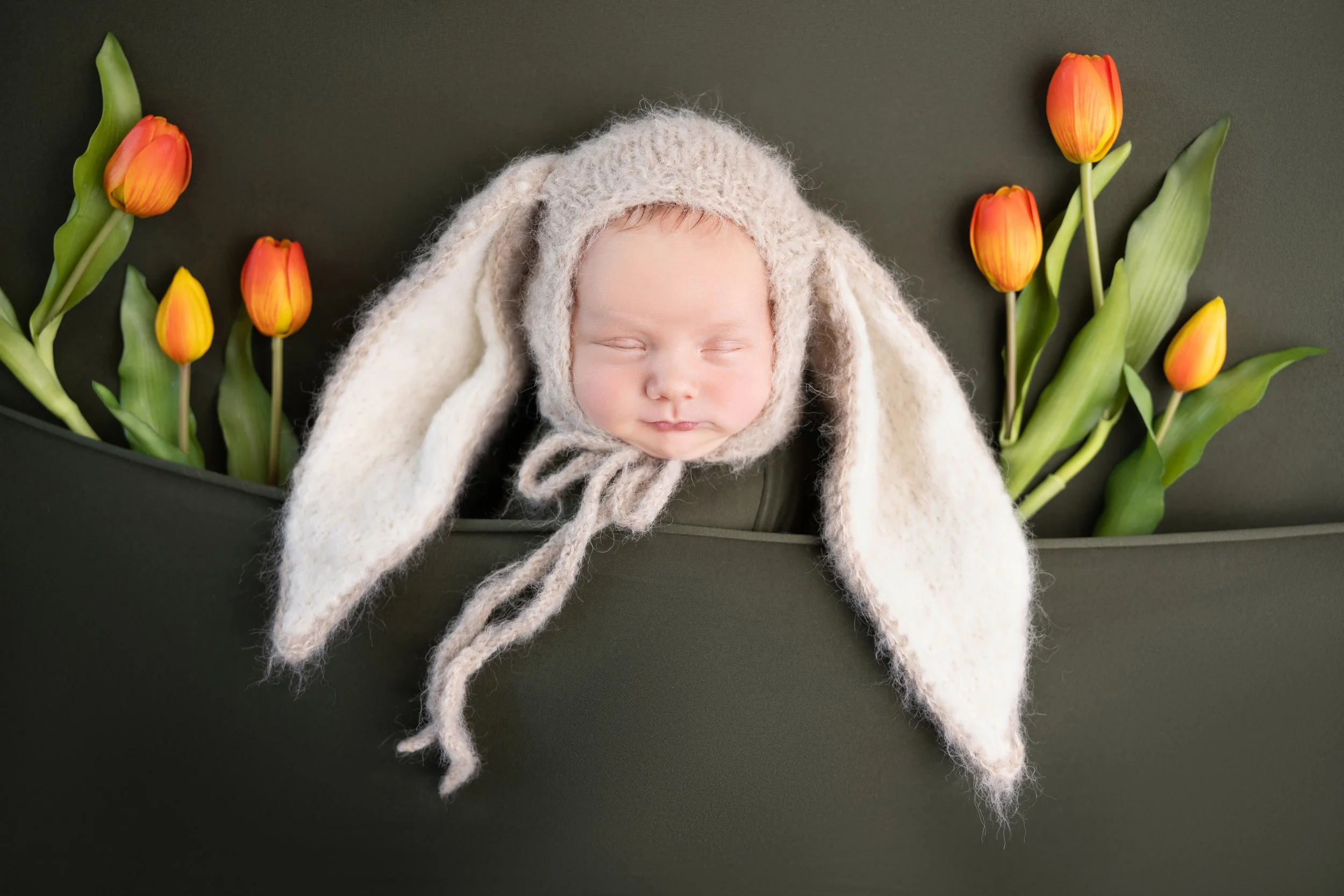 A sleeping baby wearing a bunny hat with long ears, lying amongst yellow and orange tulips on a dark background.