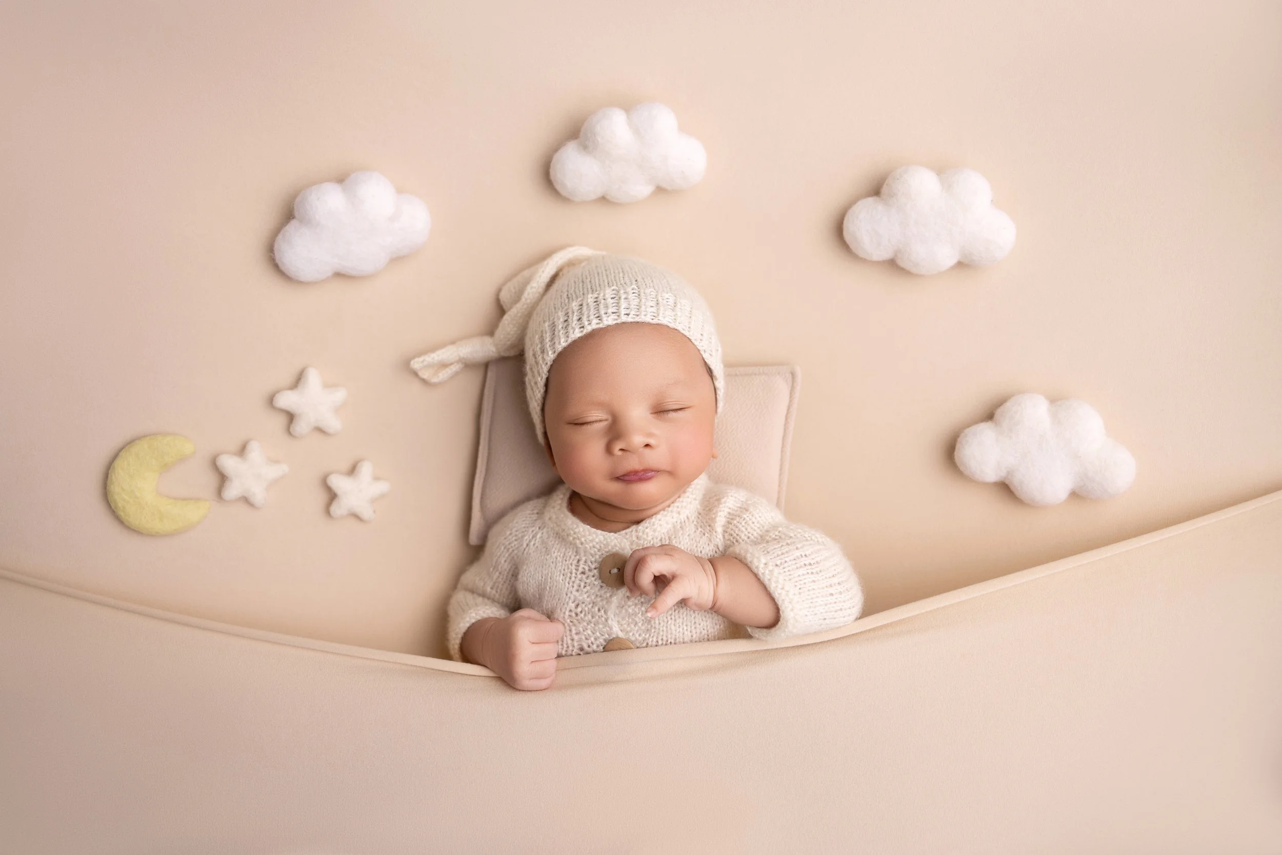 A baby with closed eyes lying in bed surrounded by felt clouds, stars, and a moon decoration on a beige background.