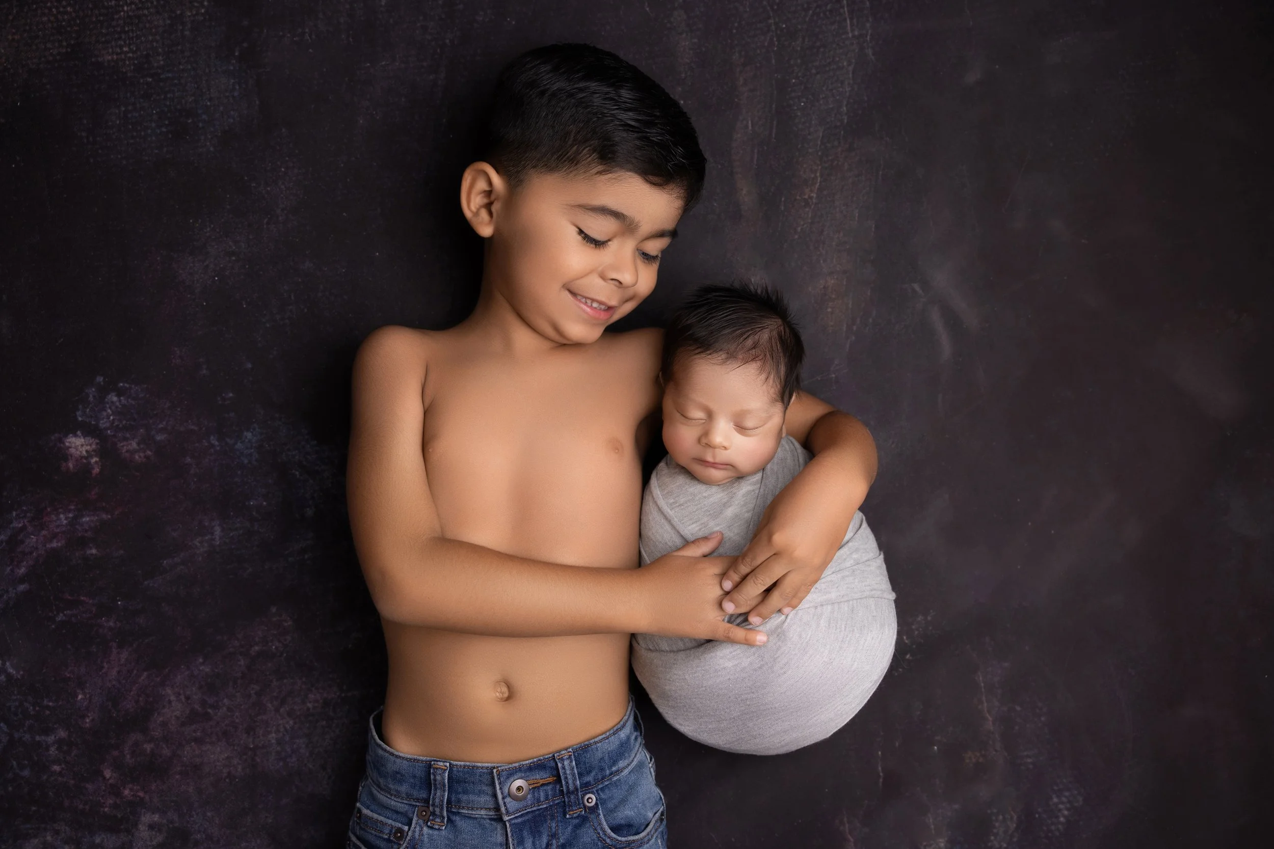 Young boy lying shirtless on a dark textured surface, holding a newborn baby snugly in his arms. Both appear peaceful with the older boy smiling softly.