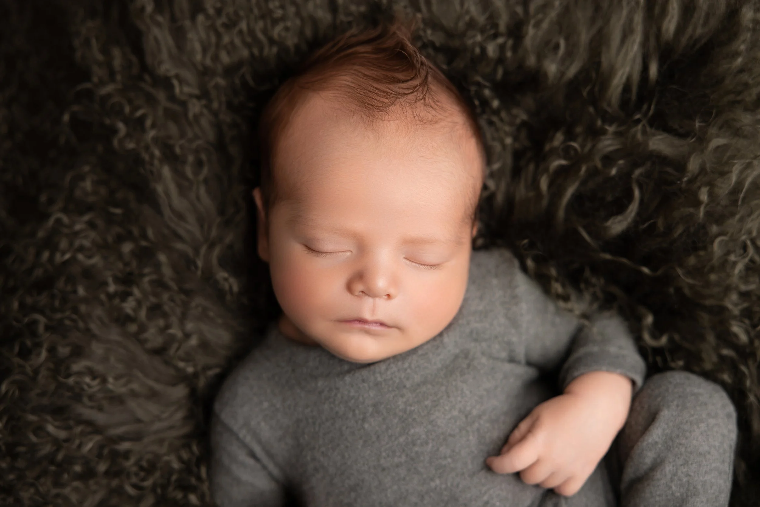 A sleeping baby with closed eyes, lying on a fluffy dark blanket, wearing a gray outfit.