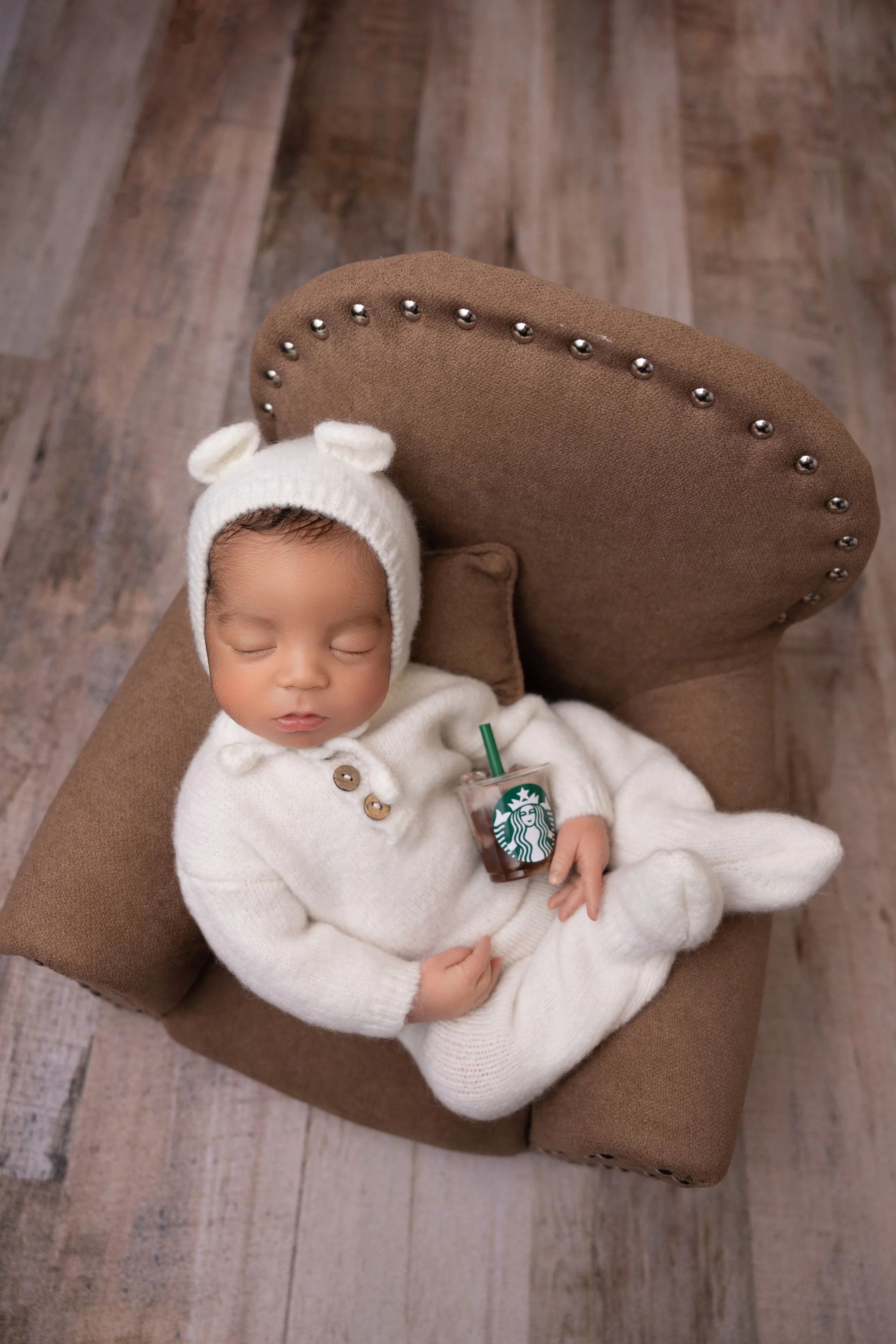 A sleeping baby in a cozy white outfit and hat with bear ears, lying on a small armchair and holding a Starbucks iced coffee.