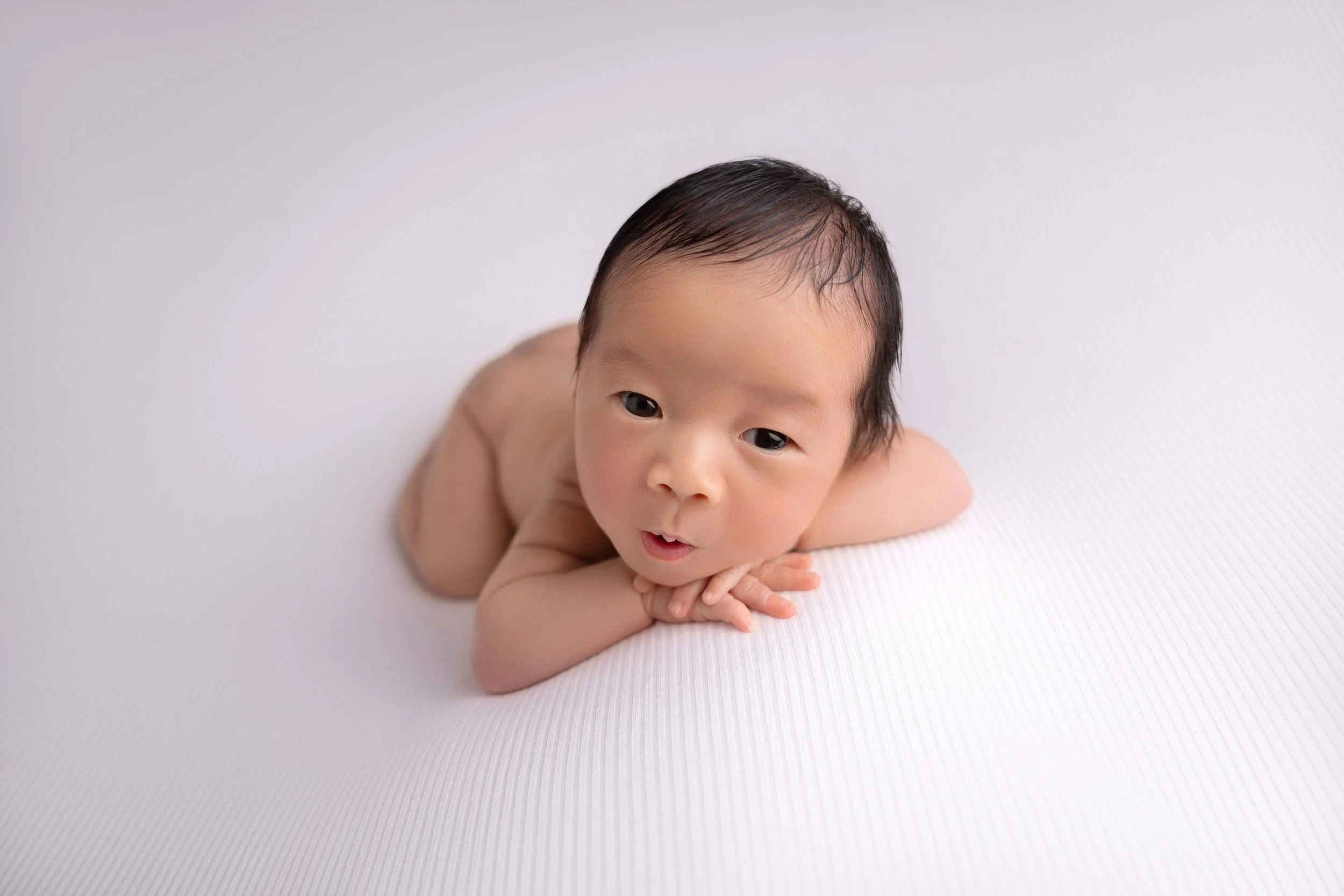 A naked baby lying on a white textured surface, looking at the camera with head resting on hands.