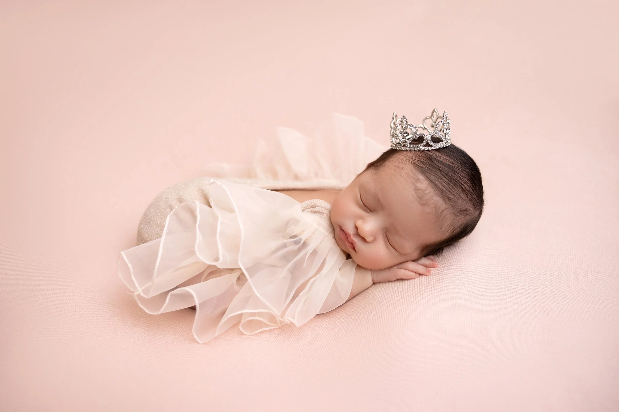 A sleeping baby with a tiara, dressed in a light-colored dress, lying on a pink background.