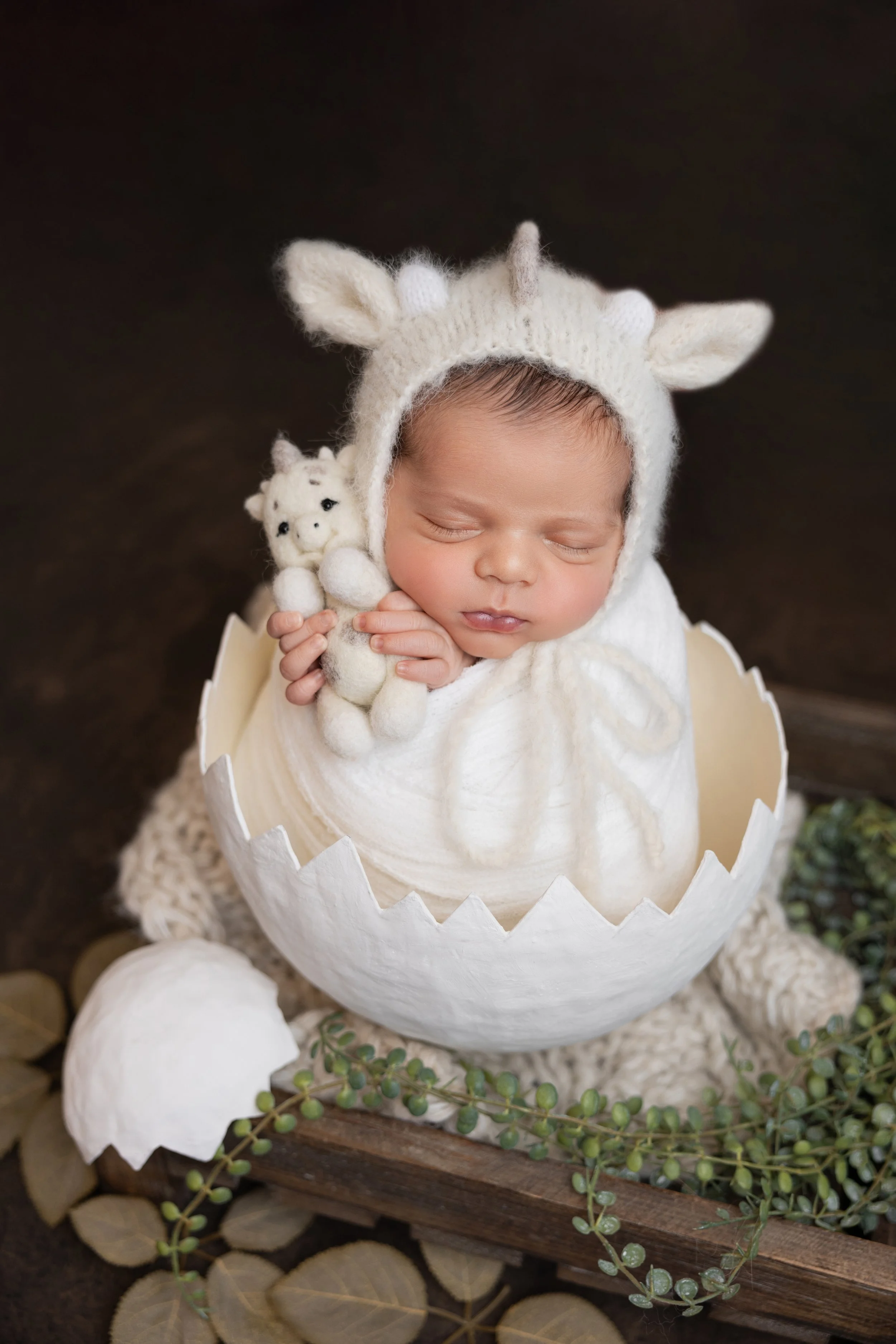 Adorable sleeping newborn baby wearing a white knitted unicorn hat, nestled inside a white eggshell prop, surrounded by greenery and a wooden tray.