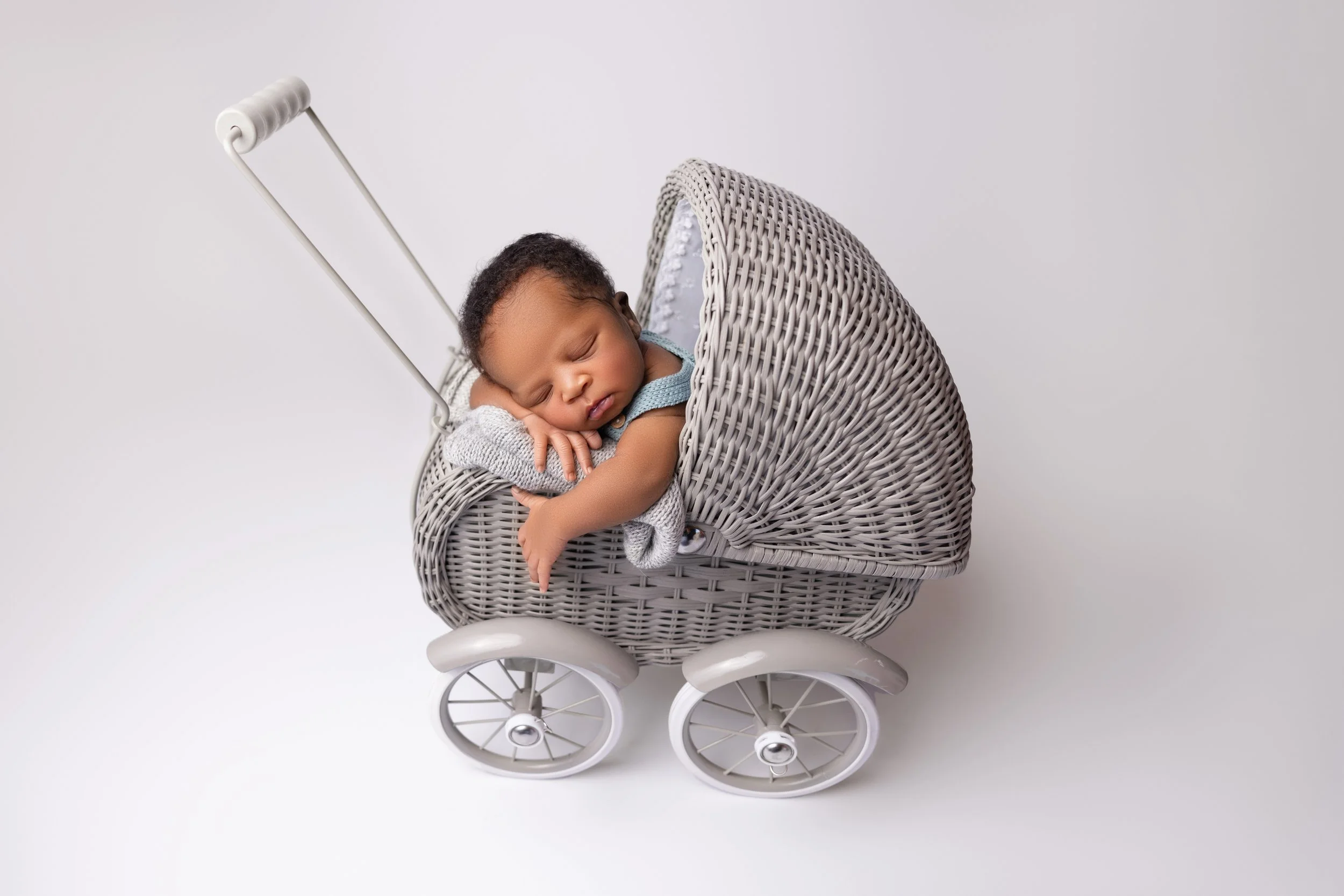 A sleeping baby with curly hair lying in a woven gray wicker toy stroller, resting with their arms crossed and head on their hands, against a plain white background.