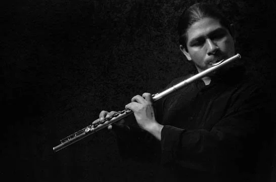 Black and white photo of a young man playing a flute, with a dark background.