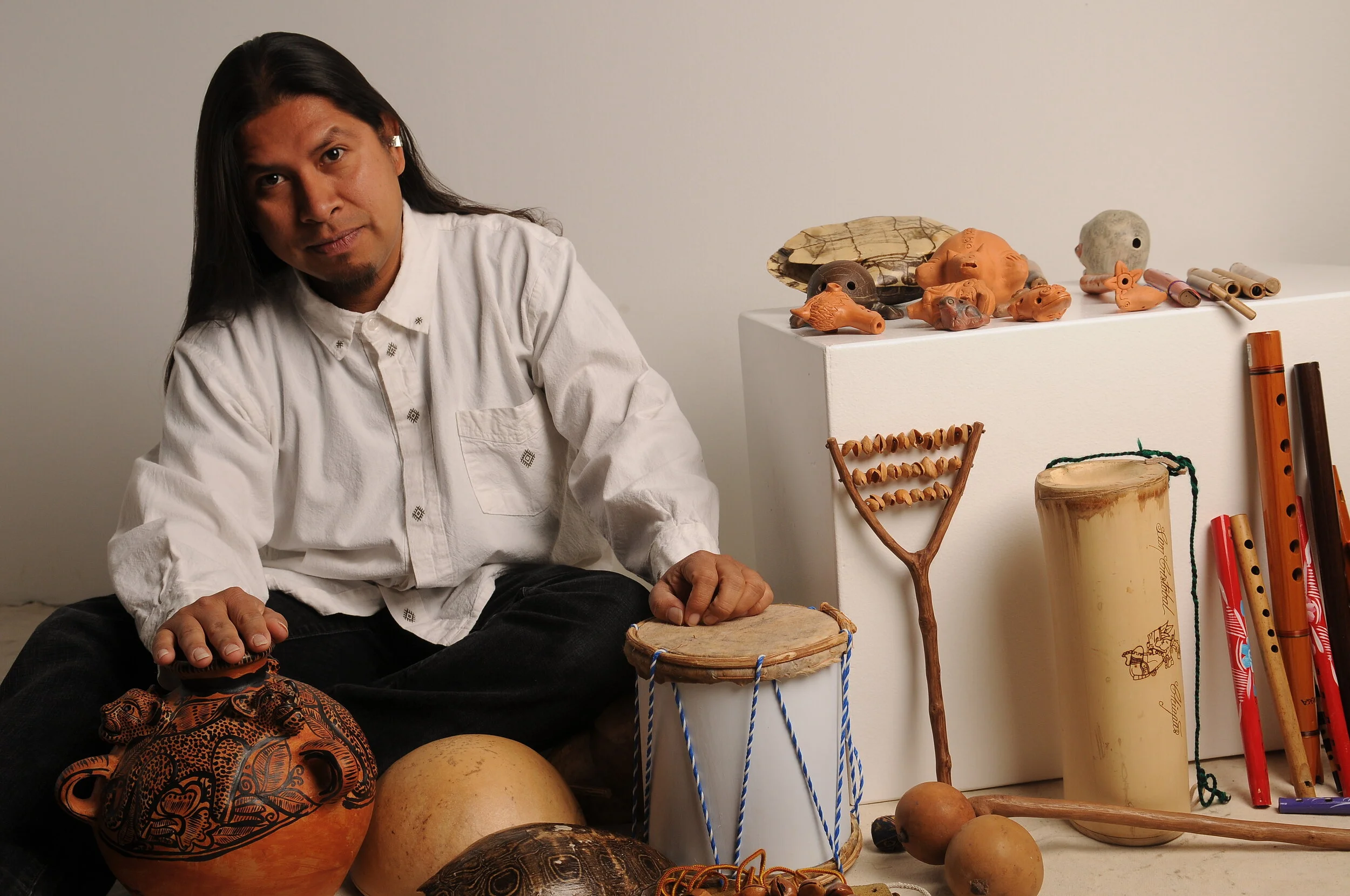 A man sitting among various traditional and indigenous musical instruments and artifacts, including drums, clay figurines, wooden sticks, and carved objects, with a plain white background.