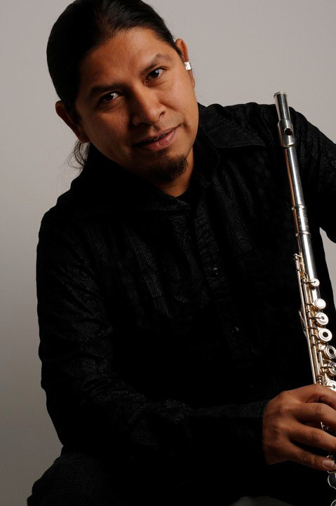 A man with long dark hair, wearing a black shirt, holding a silver flute, sitting against a plain white background.