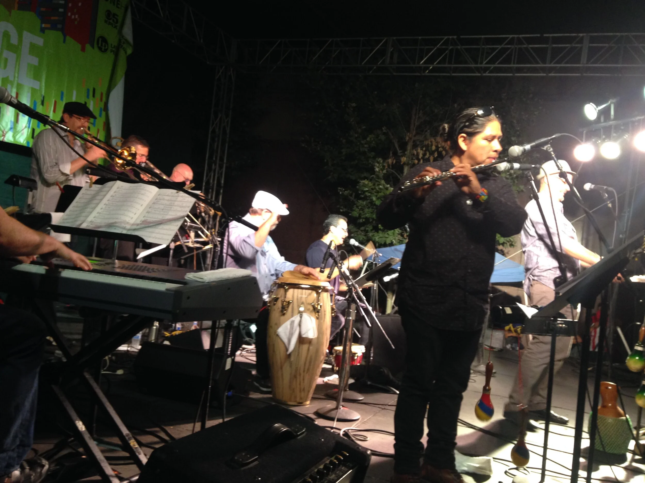 A band performing on stage at night, with musicians playing various instruments including flute, keyboard, drums, and percussion, under bright stage lights.