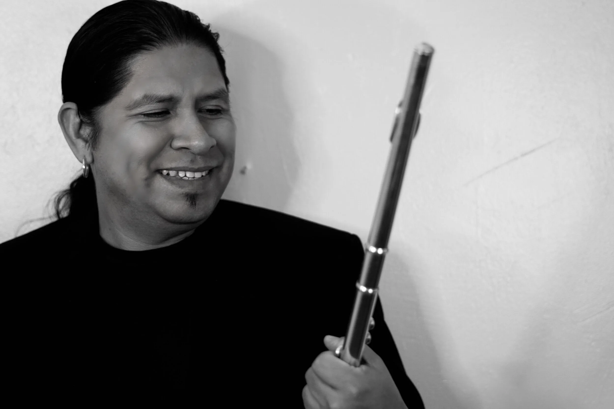 A person with long dark hair, earrings, and a goatee smiling and looking at a guitar neck.