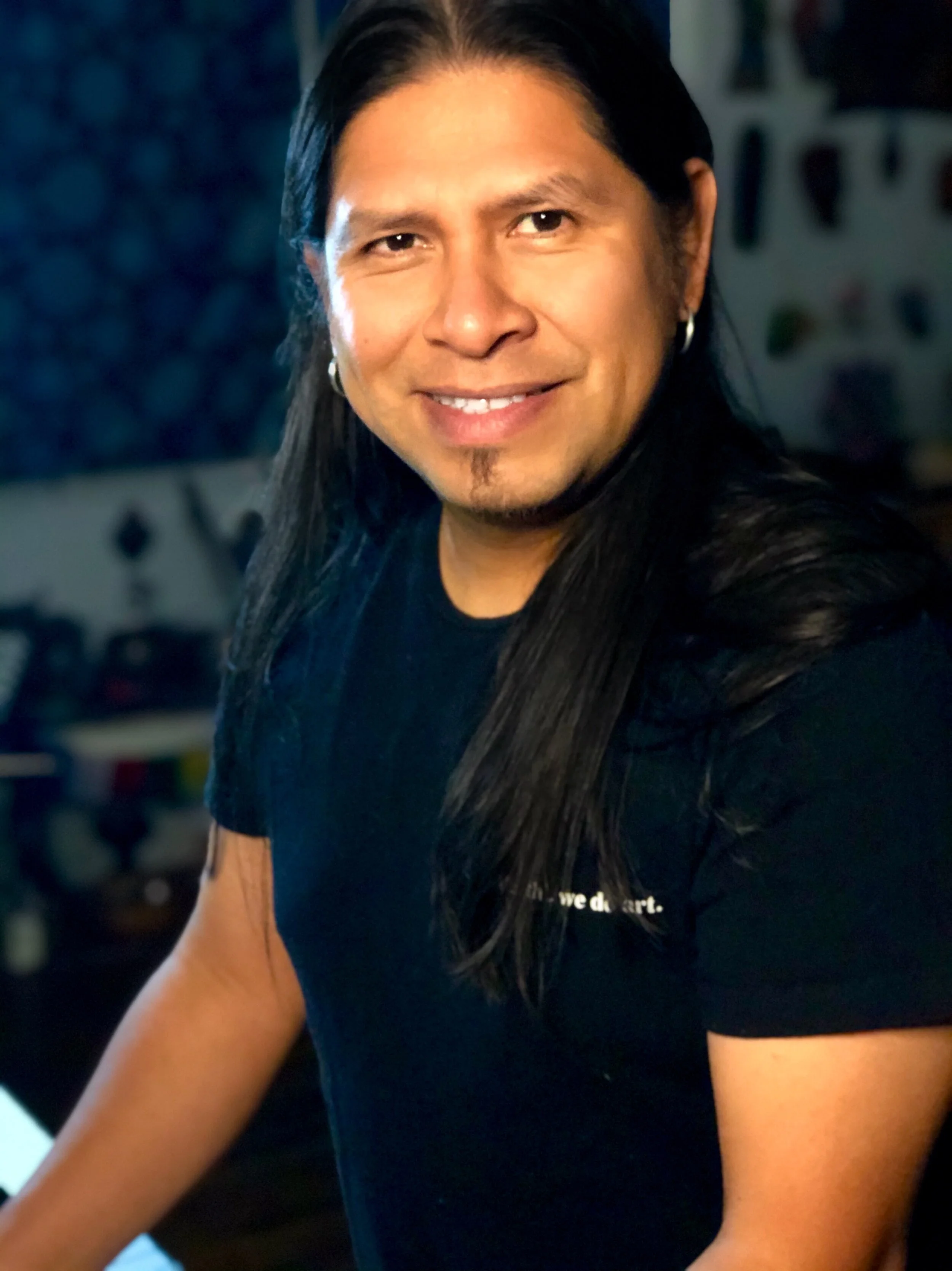 A man with long dark hair, wearing a black T-shirt, smiling at the camera, with a colorful blurred background.