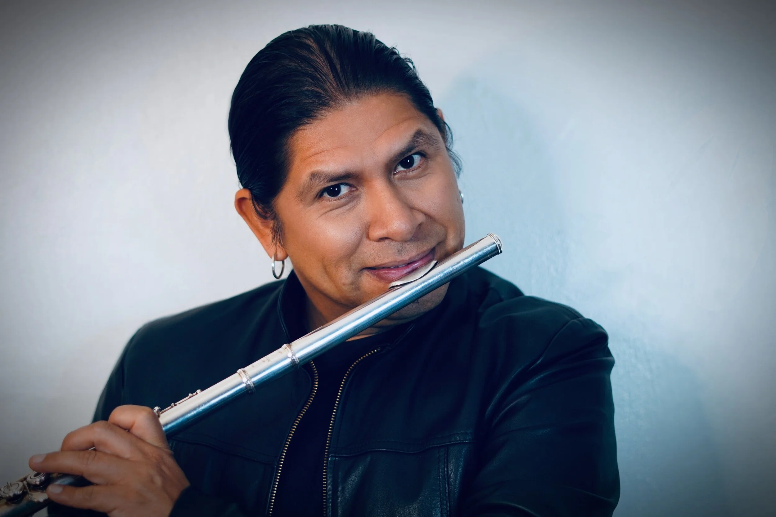 A man with dark hair and earrings playing a silver flute, wearing a black leather jacket against a plain background.