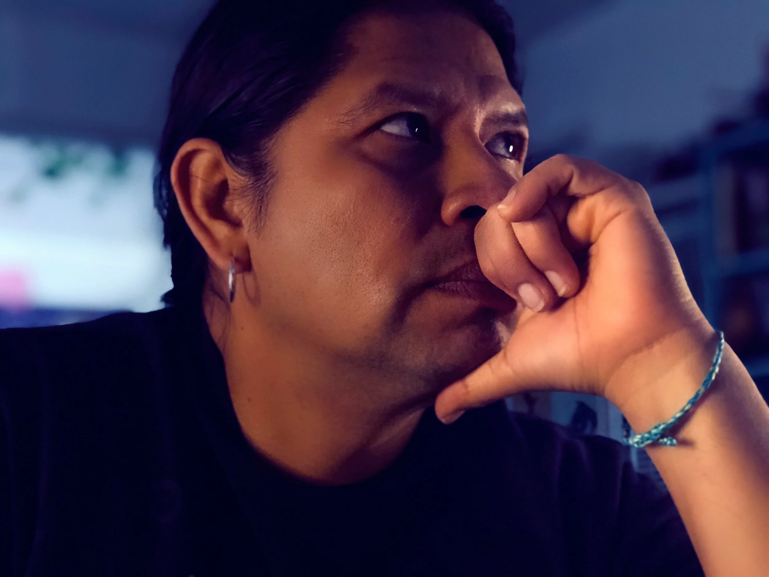 Close-up of a person with black hair, dark skin, and a hoop earring, resting their face on their hand with a contemplative expression in a dimly lit room.