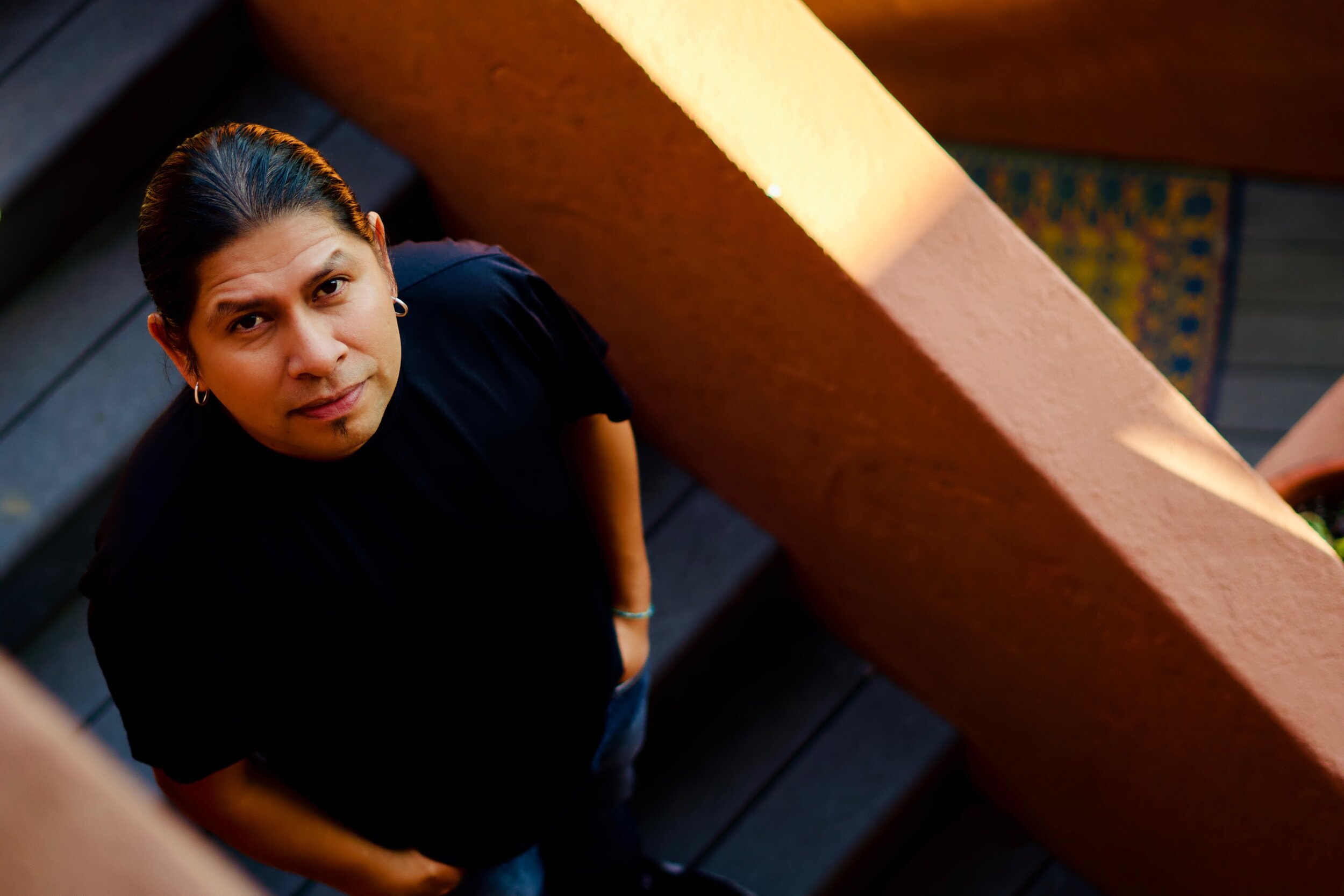 A woman with long dark hair, wearing earrings and a black shirt, looking up at the camera from a staircase landing, with a peach wall and patterned tile in the background.
