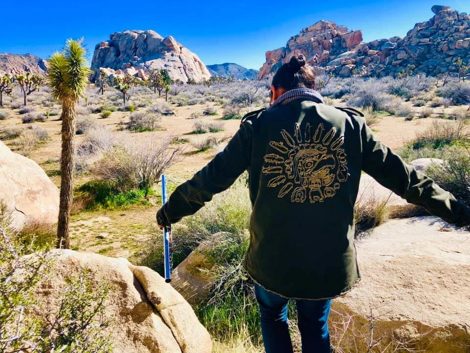 A person standing on a rock in a desert landscape with rocky hills in the background, holding a measuring stick, wearing a jacket with a sun-themed design on the back.