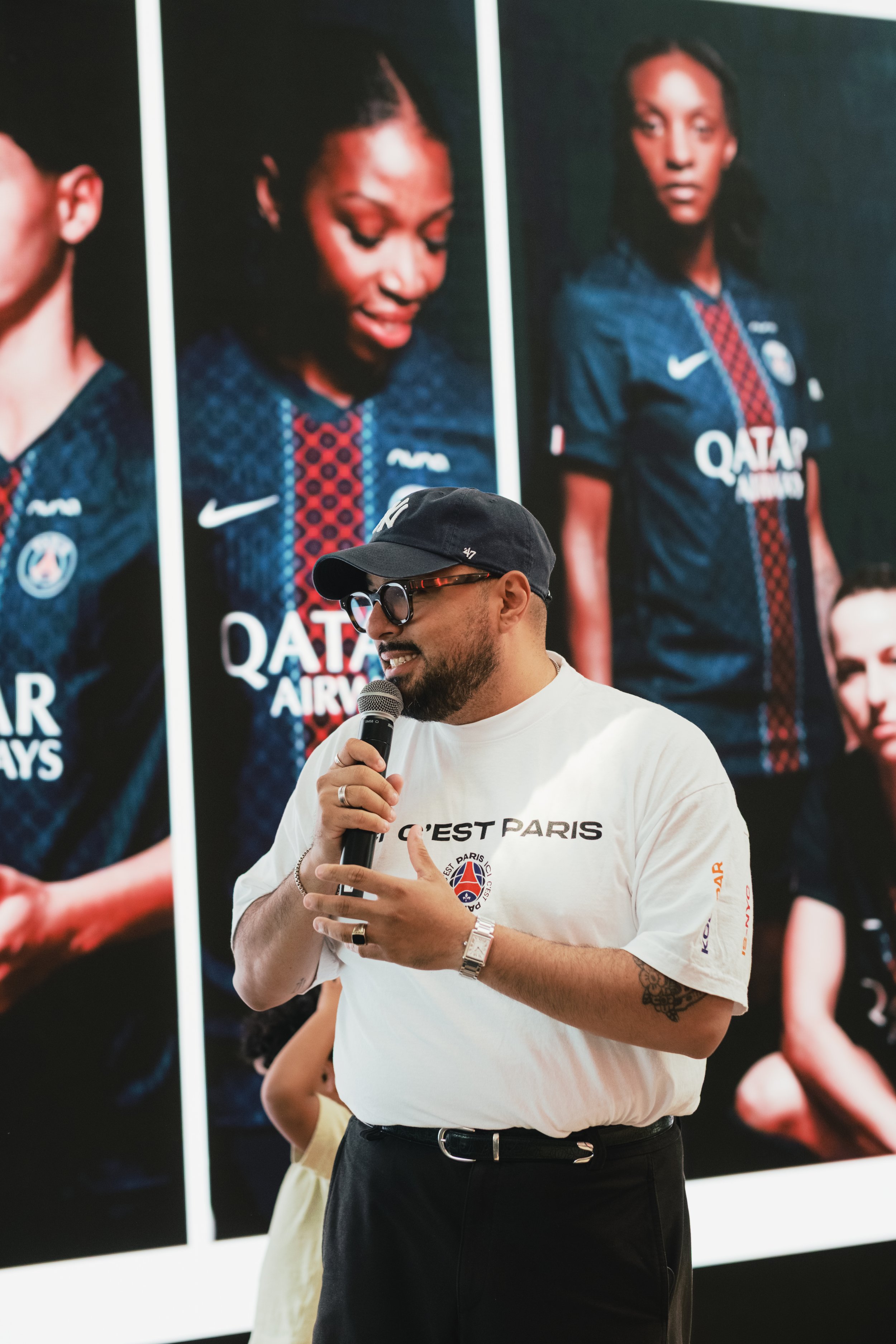 A man wearing a black cap, glasses, and a white T-shirt with a logo, holding a microphone and speaking, standing in front of a large poster of two women in athletic jerseys.