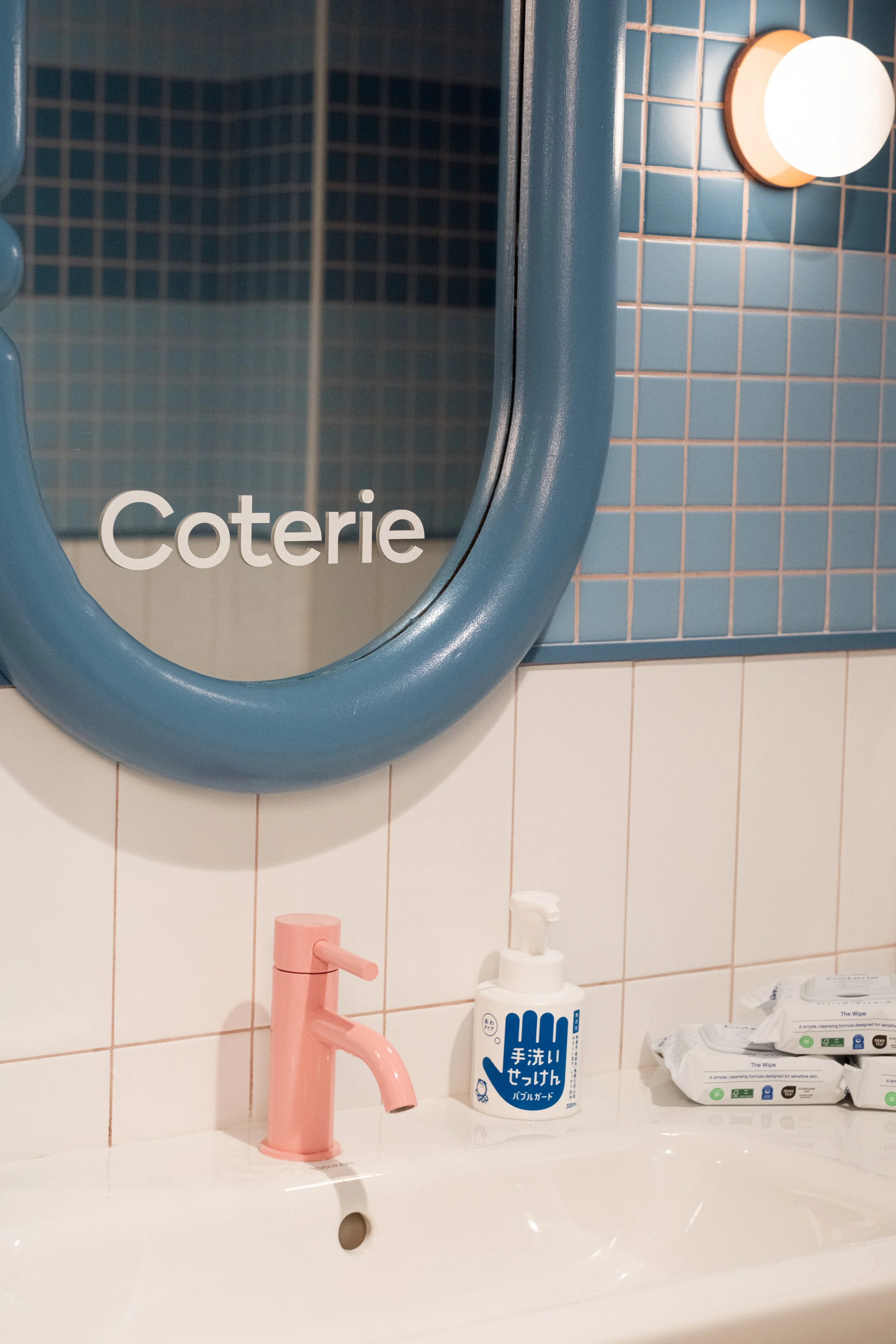 Bathroom sink with a pink faucet, a soap dispenser, and a mirror labeled 'Coterie' on a tiled wall with a blue and white color scheme.