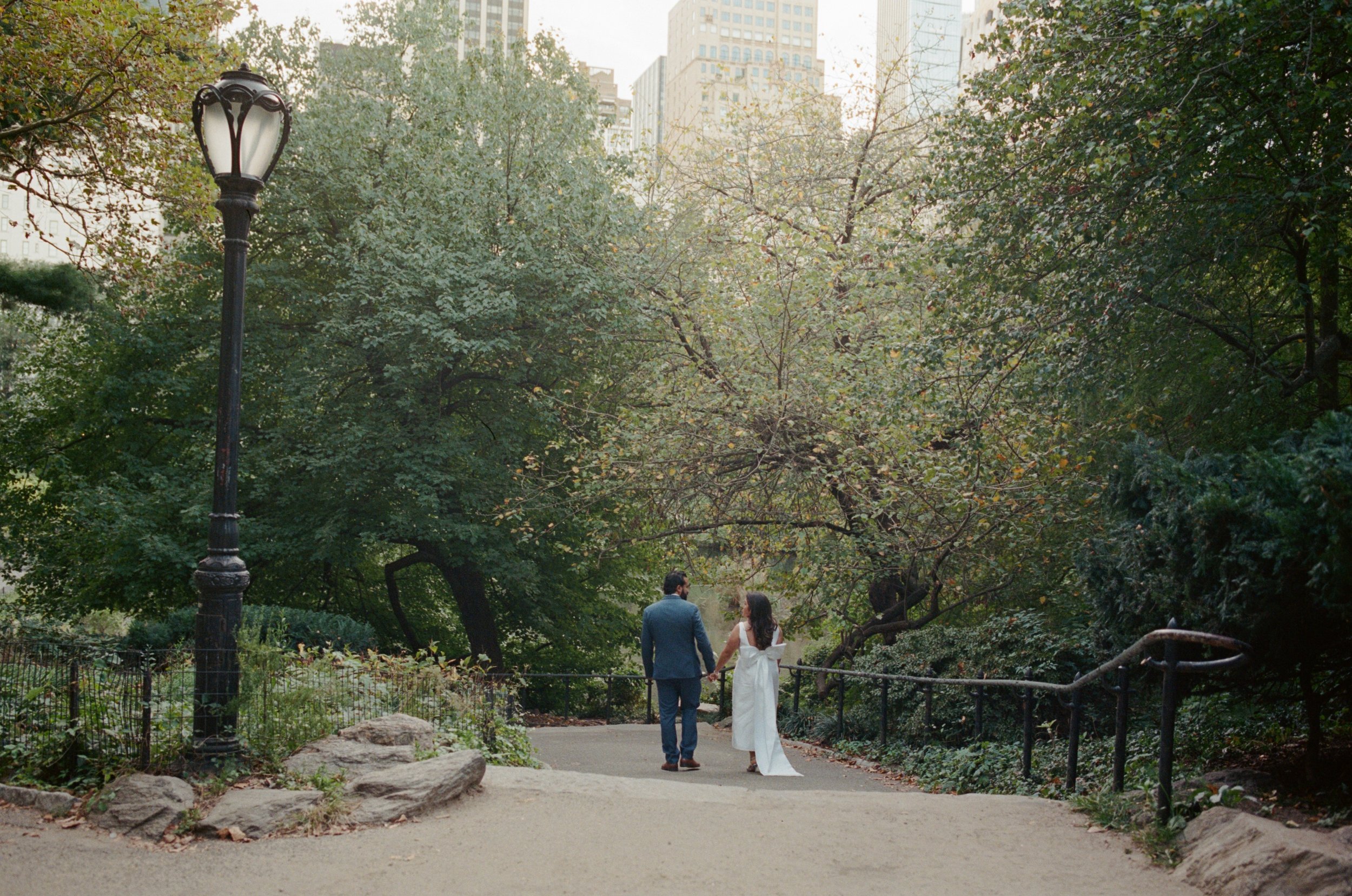 A man and woman dressed in wedding attire walking hand in hand through a park with green trees and city buildings in the background.